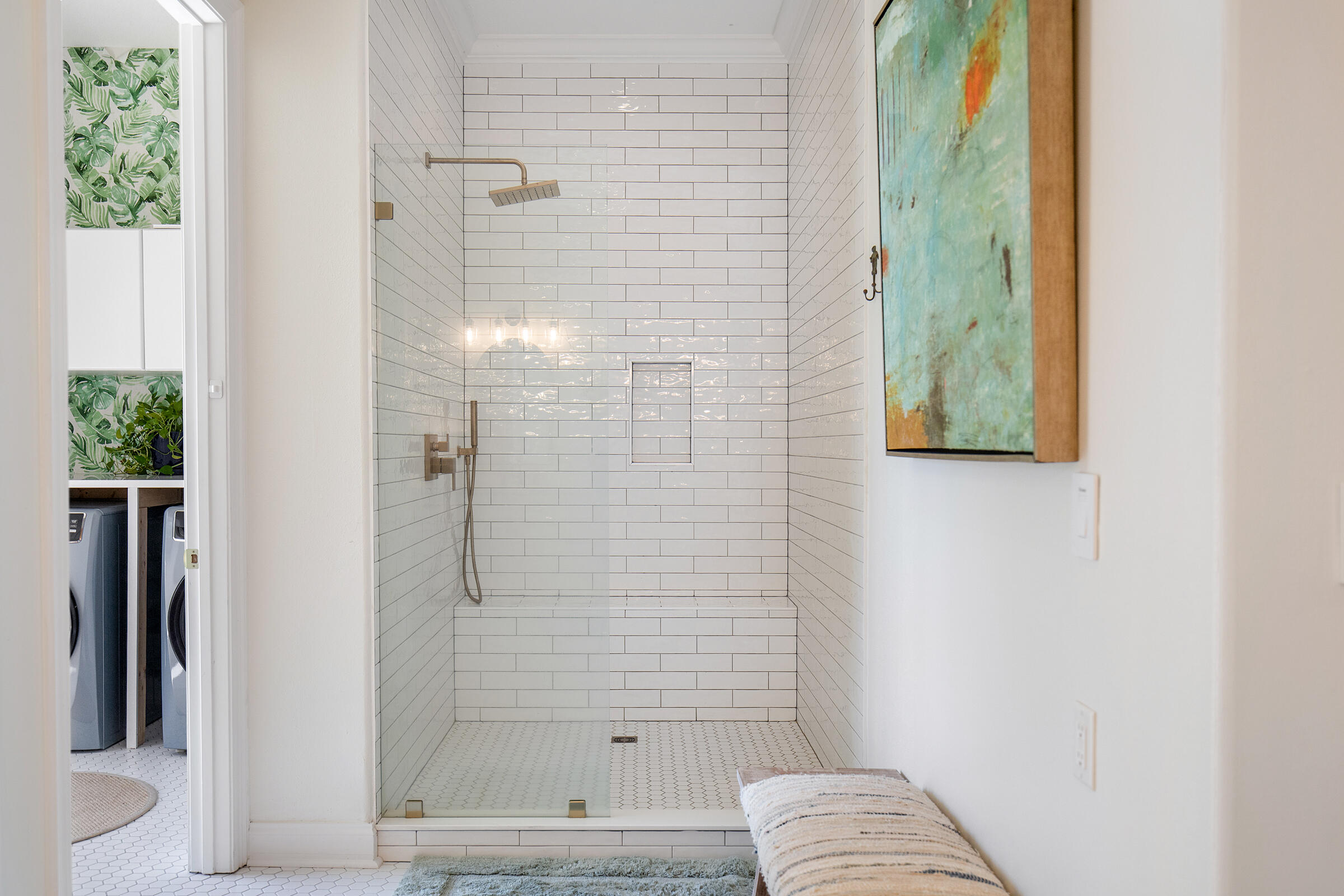 Walk-in shower with white subway tile, brass fixtures, and a glass panel beside a laundry area.