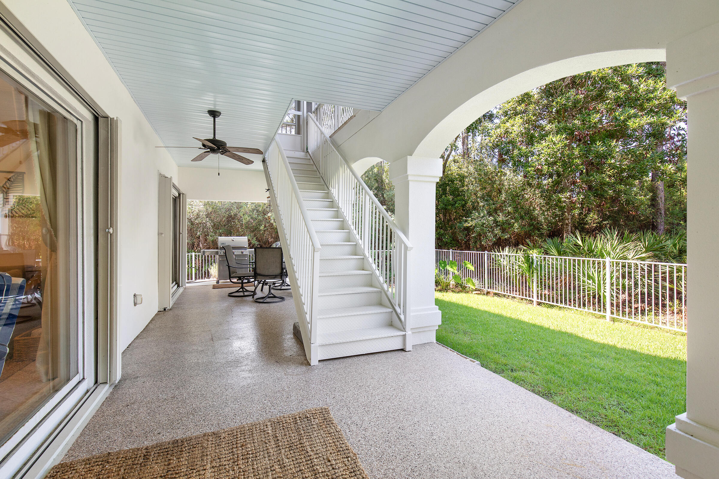 Covered patio with white staircase, ceiling fan, and fenced lawn bordered by trees.