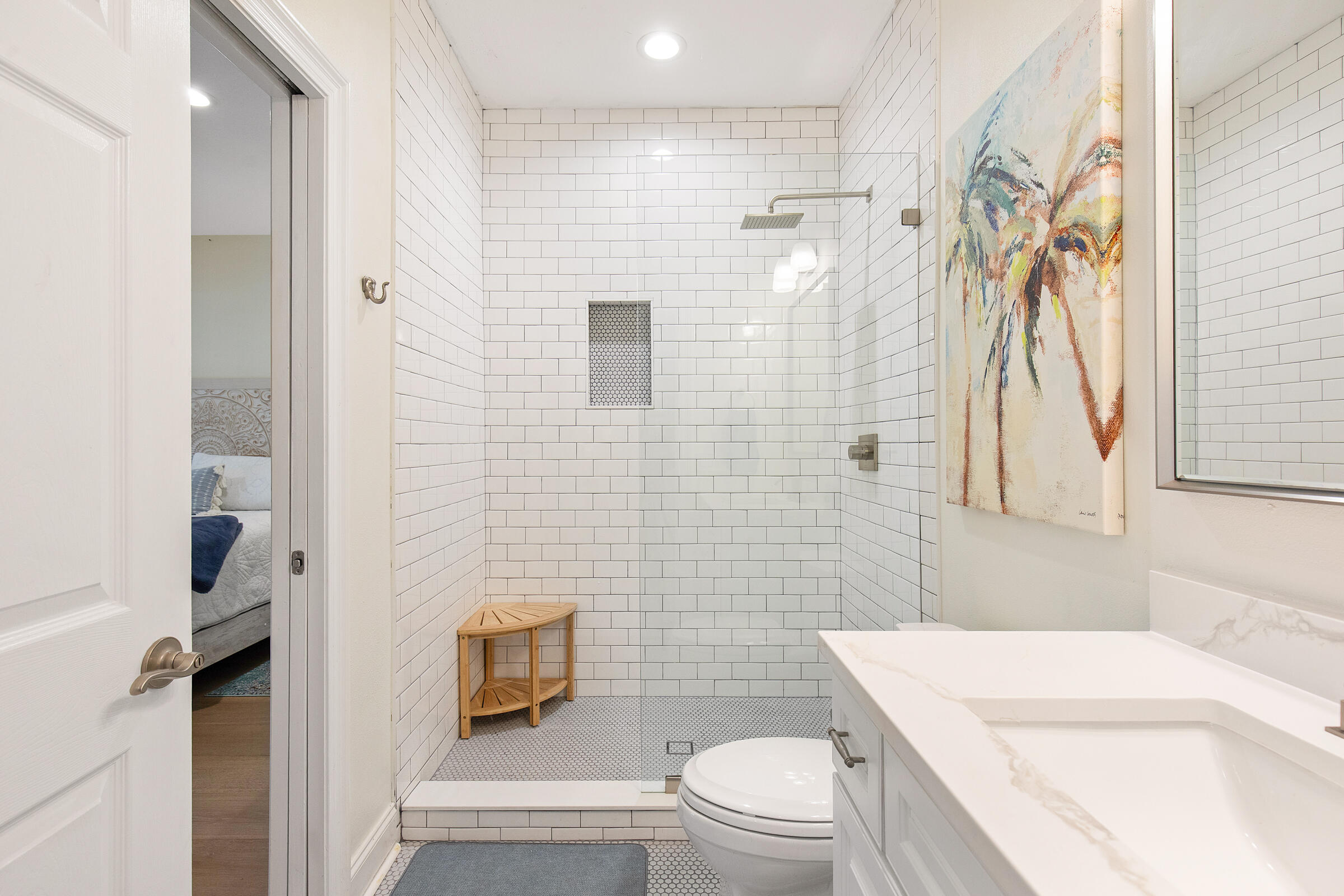 Bathroom with a white subway-tile walk-in shower, wooden corner stool, and colorful abstract artwork.