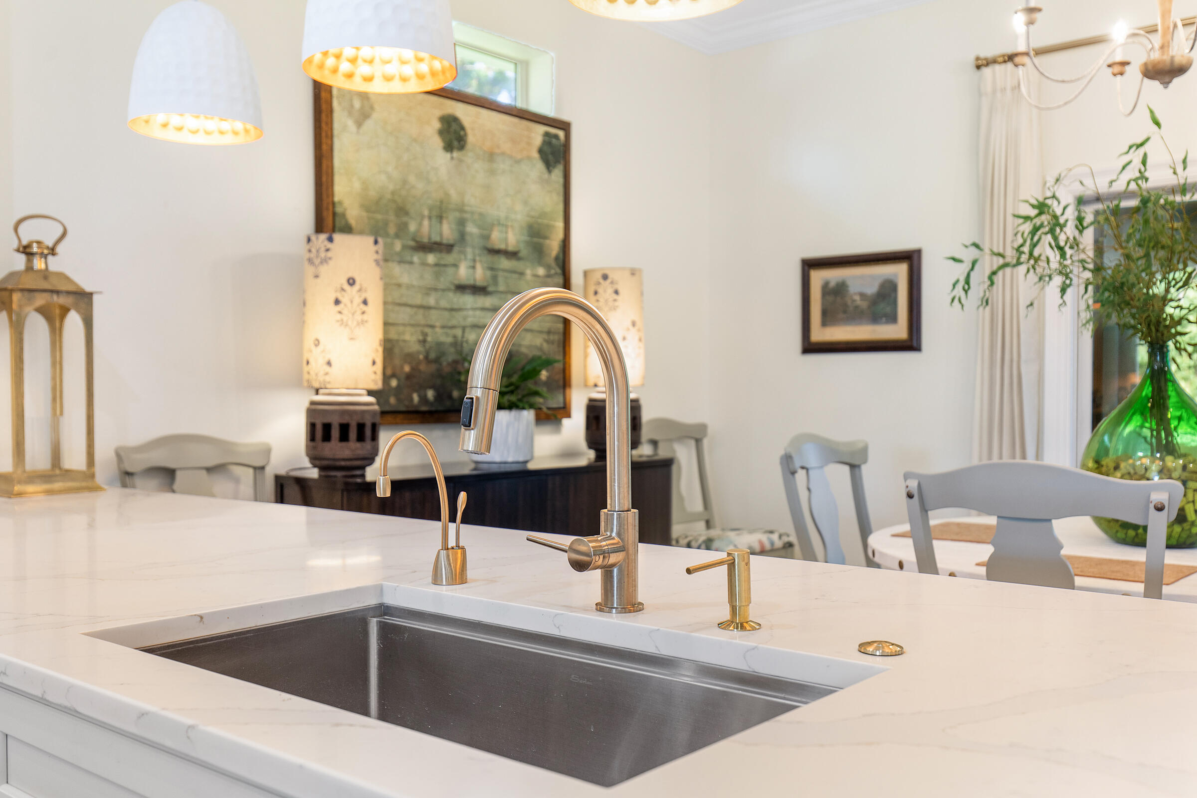 Kitchen sink with white veined countertop, brass-tone faucets, and dining area beyond.