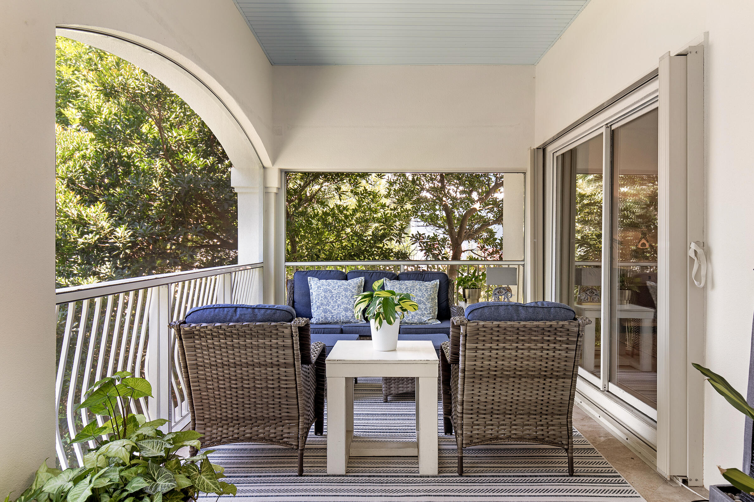 Covered porch with blue sofa, wicker chairs, striped rug, and leafy views through white railings.