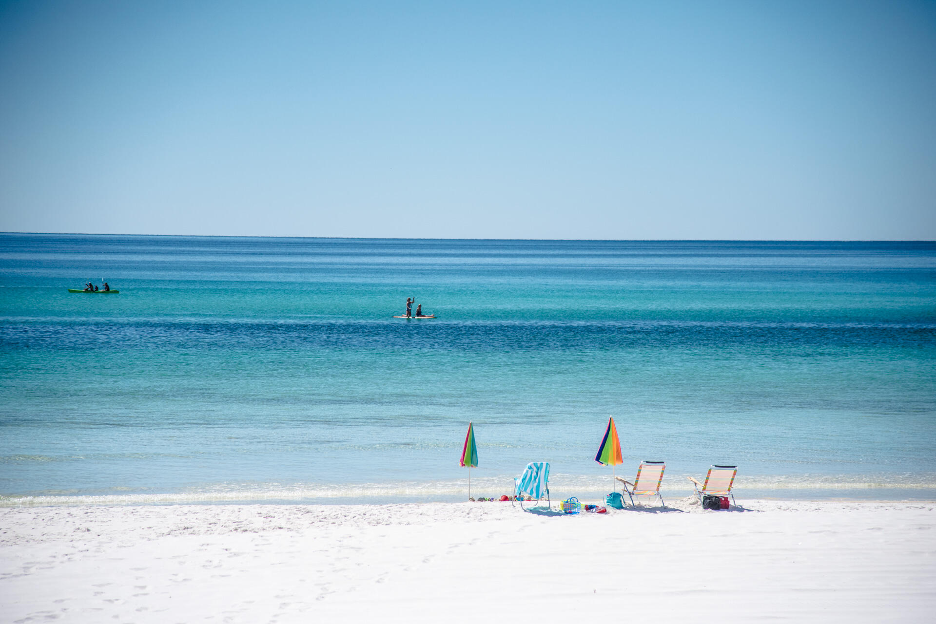 White-sand beach with colorful umbrellas and chairs, and paddleboarders on calm turquoise water.