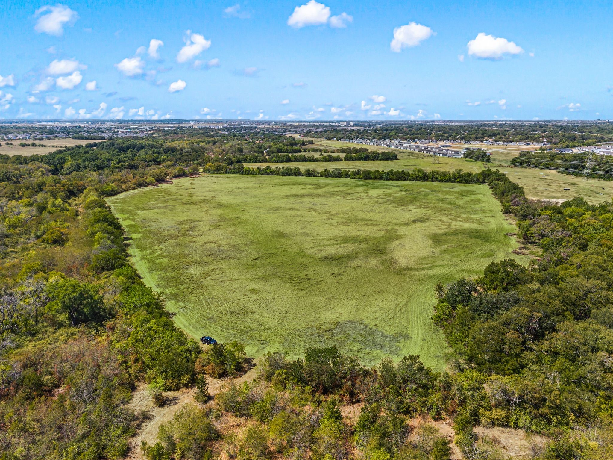 Aerial view of a broad grassy clearing bordered by woods, with homes visible in the distance.