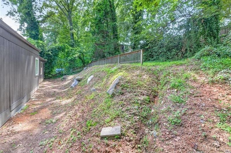Sloped wooded backyard beside a dark-sided house, with a dirt path and wooden railing fence.