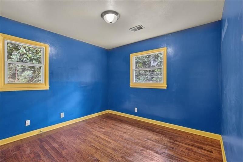Empty room with blue walls, yellow trim, reddish wood flooring, and two windows overlooking trees.