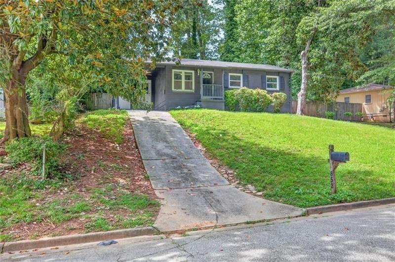 Gray single-story house behind a broad lawn, with a concrete driveway and mature trees.