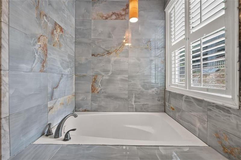 Gray marble-look tiled bathroom with a white soaking tub beneath a plantation-shuttered window.