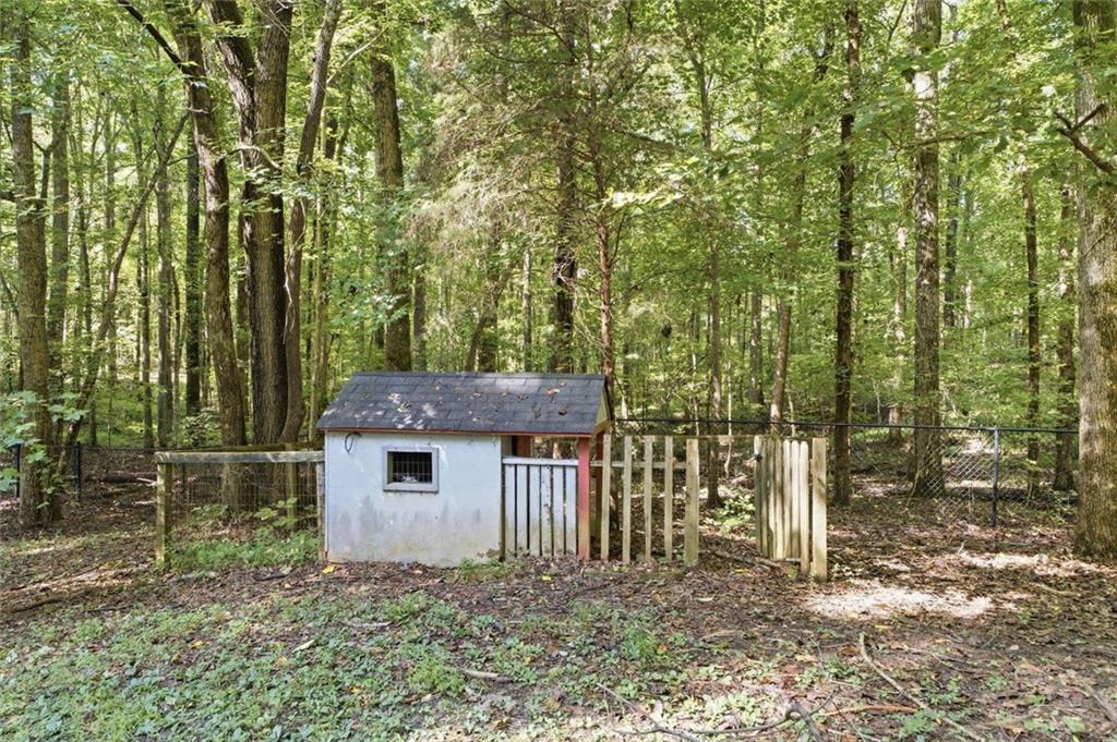 Small white kennel behind a low fence in a wooded yard surrounded by tall leafy trees.