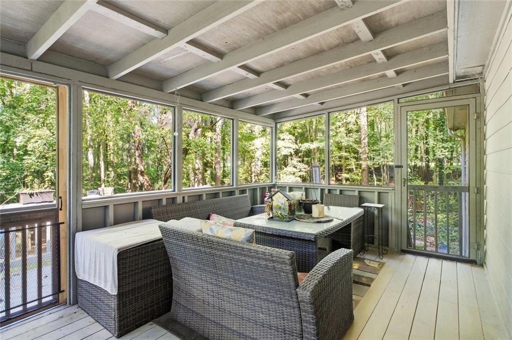 Glass-enclosed porch with wicker seating and dining table overlooking wooded surroundings.