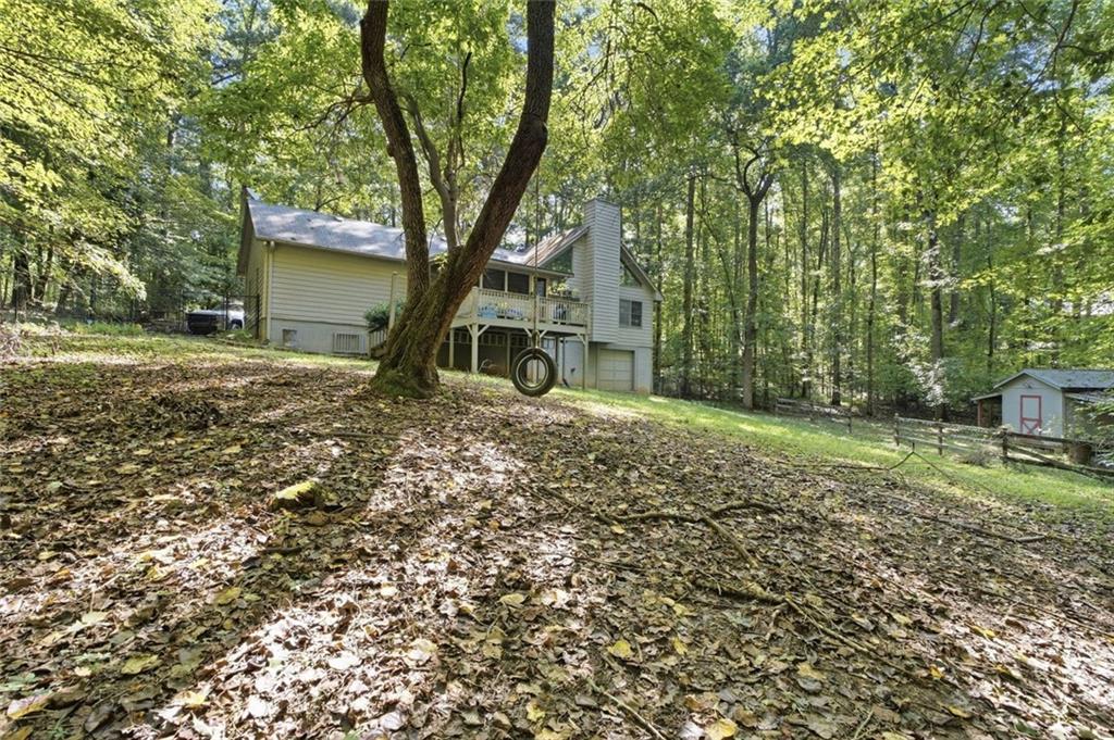 Wooded house with a raised deck, large tree, leaf-covered yard, and small outbuilding.
