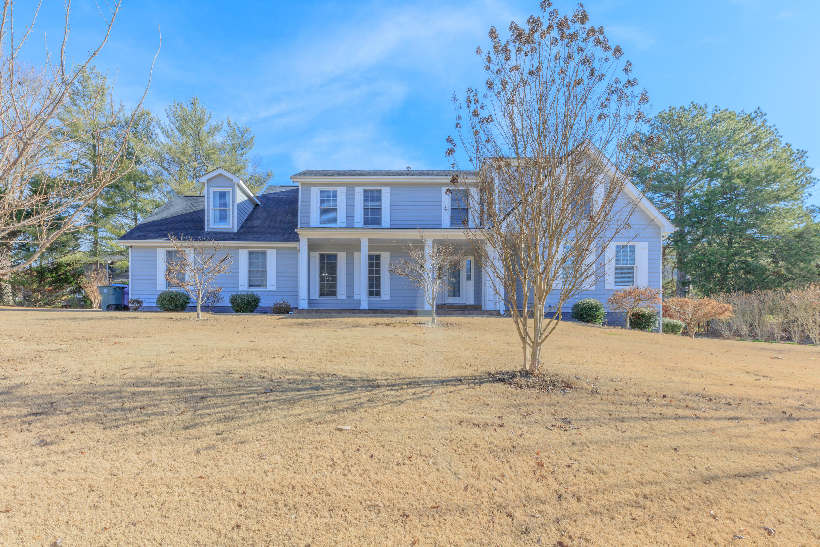 Two-story blue house with white trim and a covered porch behind a broad dormant lawn.