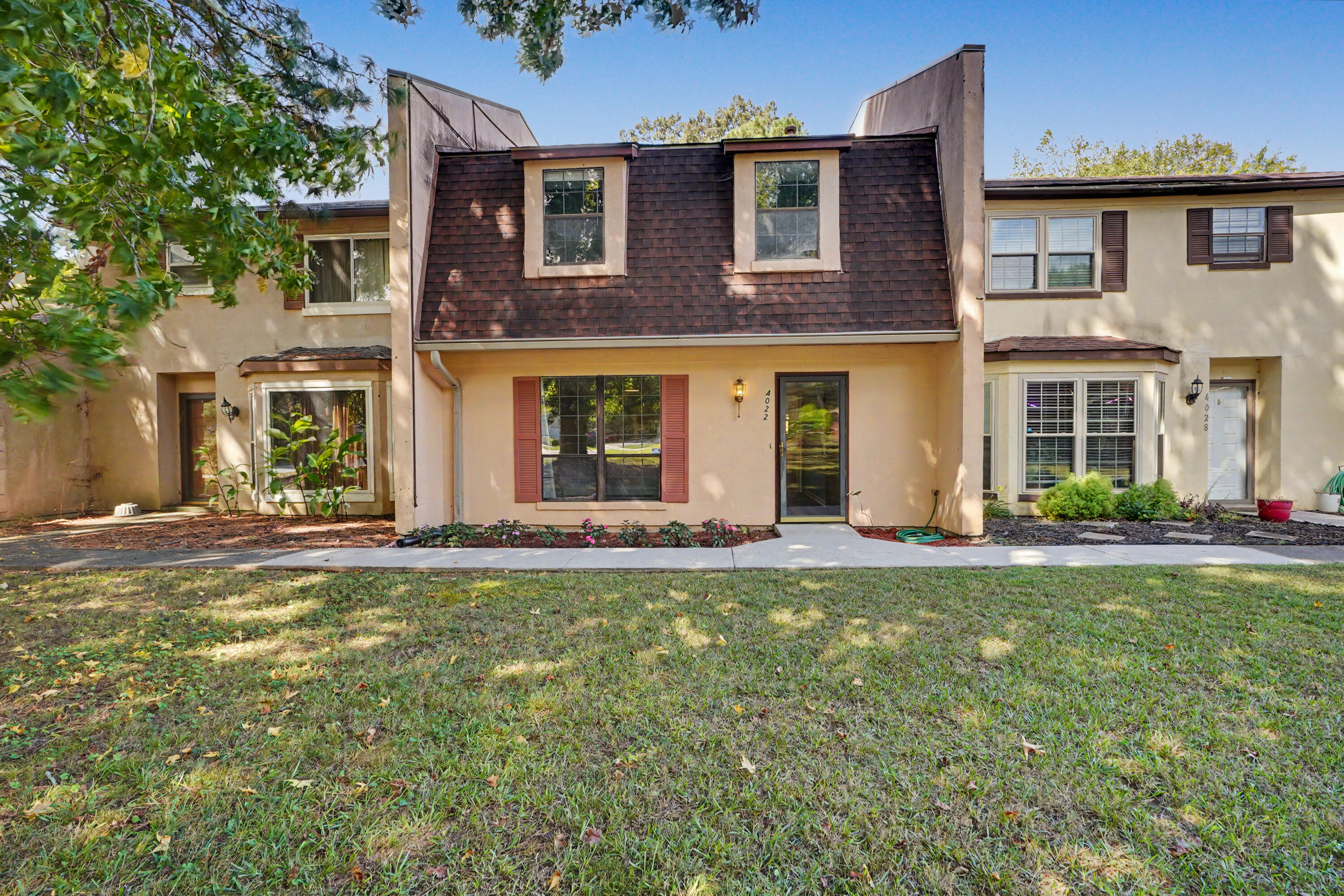 Two-story beige townhouse with a steep dark shingle roof, dormer windows, and a front lawn.