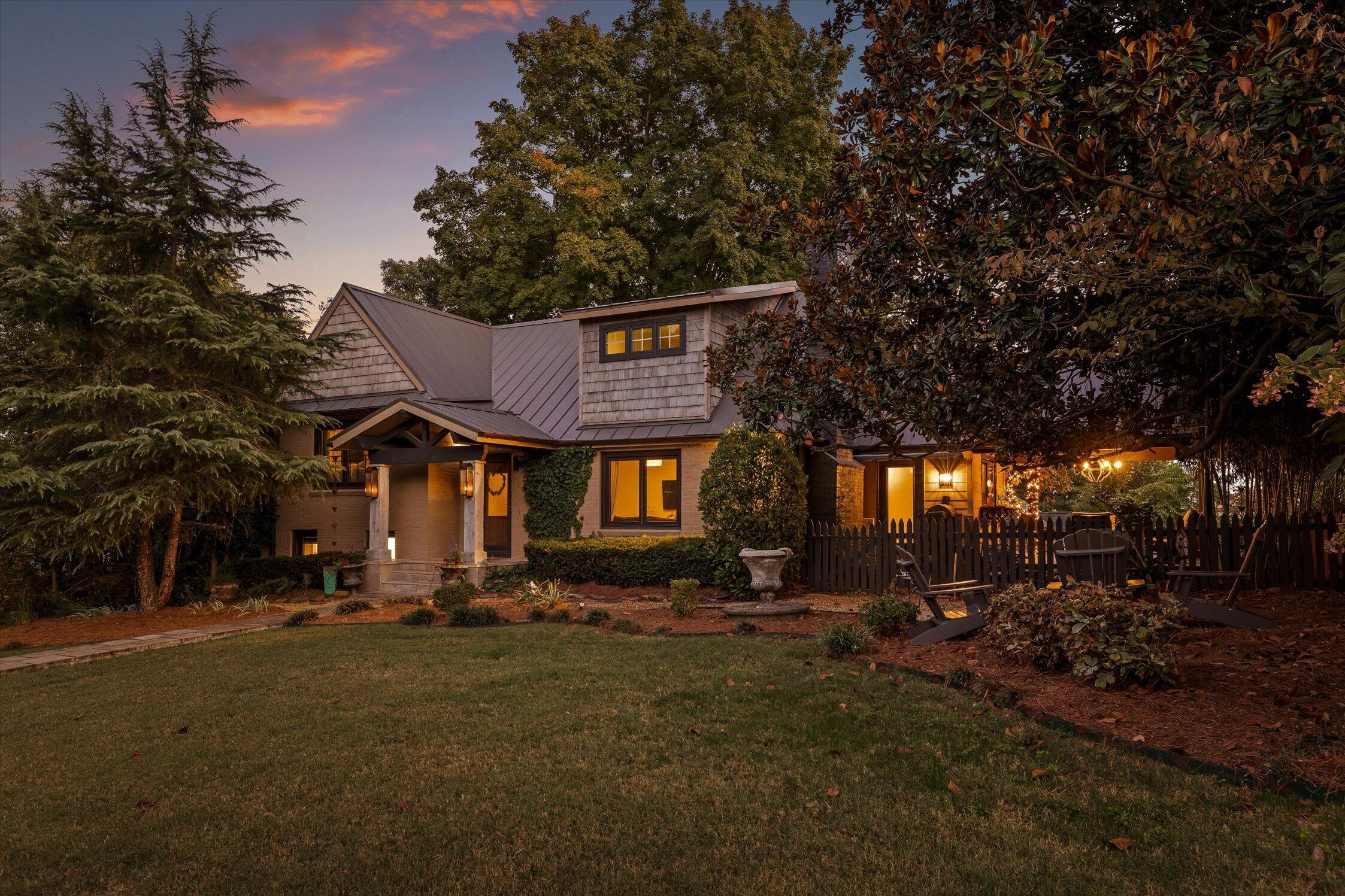 Two-story home with a metal roof, landscaped lawn, and warmly lit fenced patio at dusk.