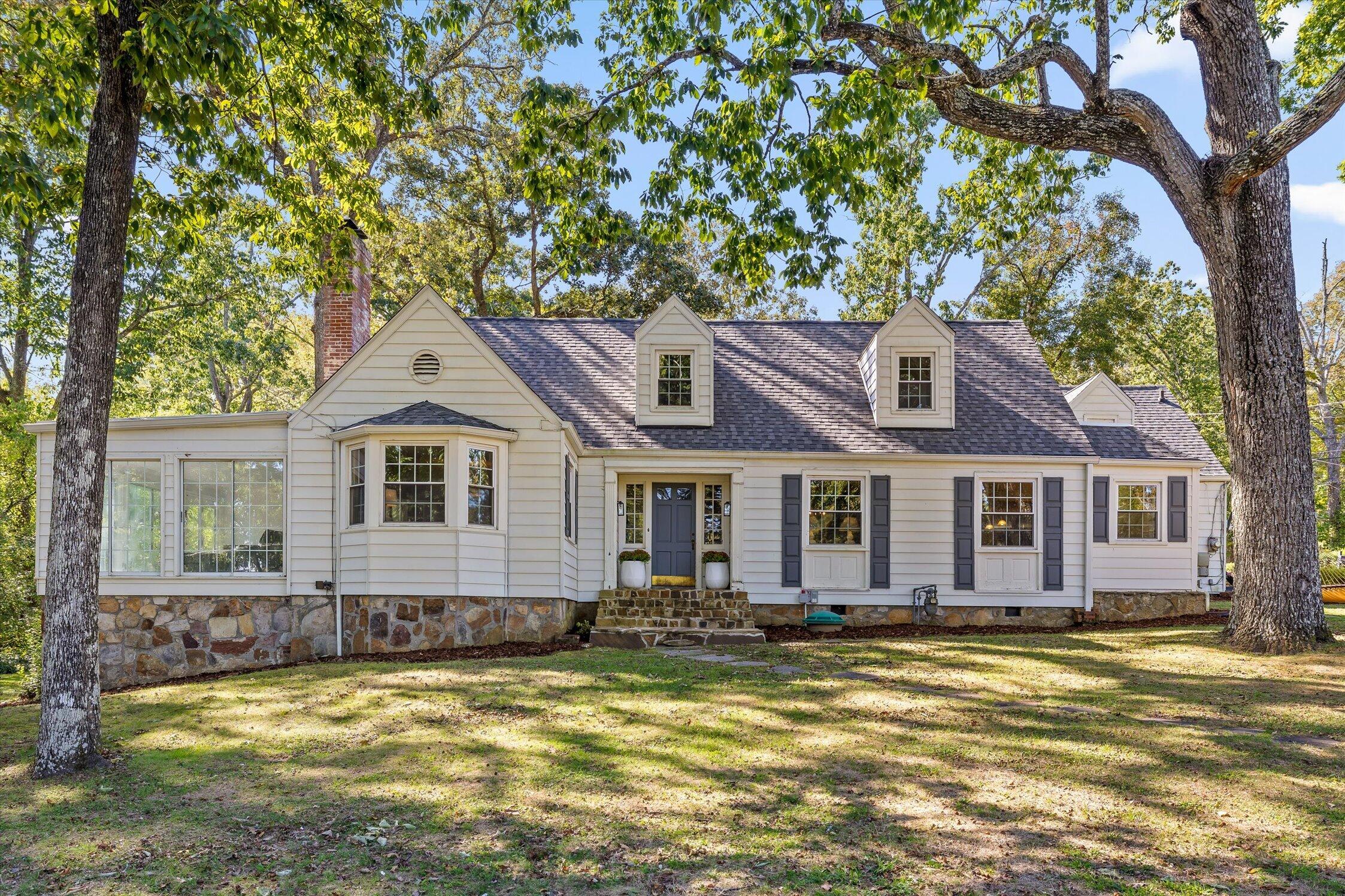 White-sided house with dormer windows, a stone foundation, and a shaded lawn beneath mature trees.