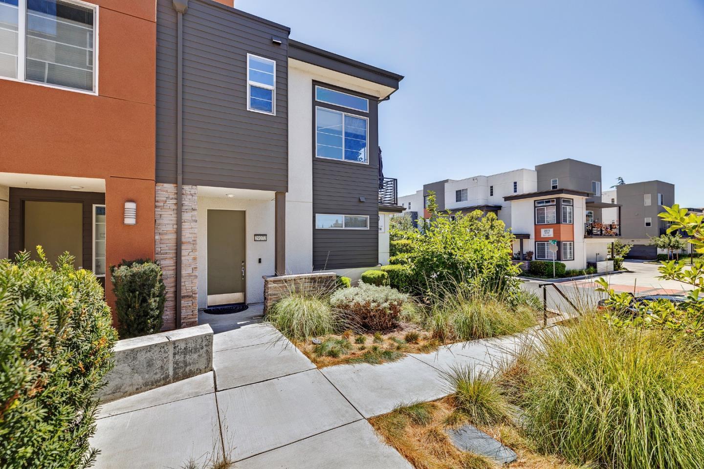 Modern townhomes with orange and gray facades beside a landscaped concrete walkway.