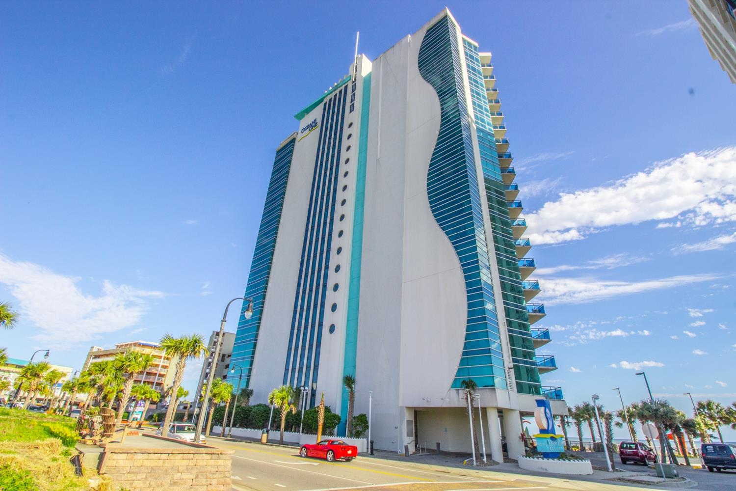 Modern white condominium tower with curved turquoise glass façades and balconies above a palm-lined street.