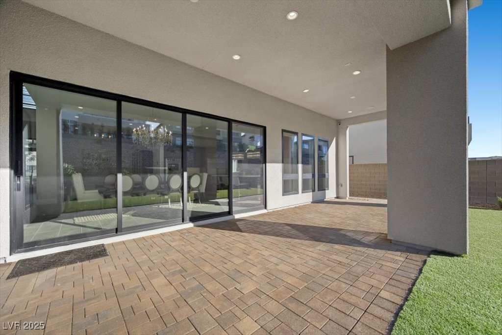 Covered backyard patio with tan pavers, broad sliding glass doors, and a strip of artificial turf.