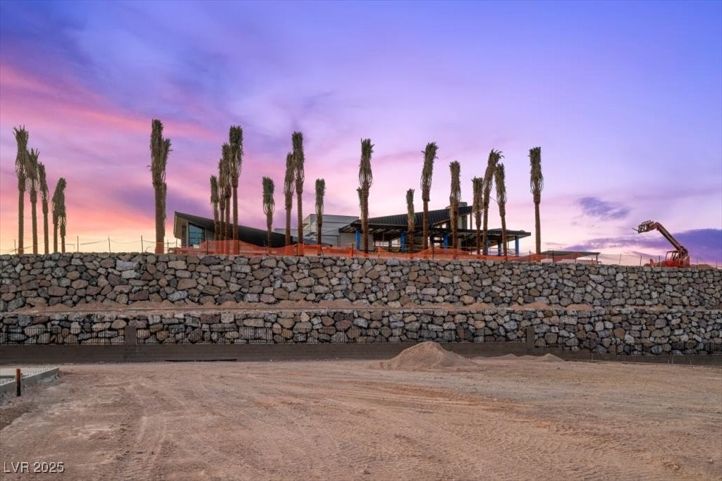 Modern home under construction behind a stone retaining wall at dusk, with tall palms.