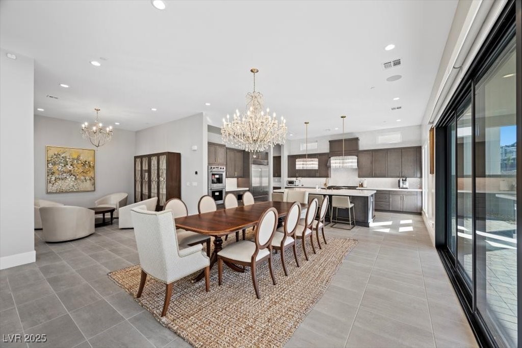 Open-plan dining room and kitchen with a long wood table, chandeliers, dark cabinetry, and sliding glass doors.