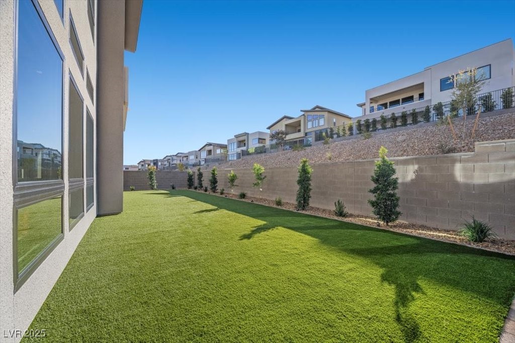 Side yard with a broad green lawn, house windows, and a block wall lined with young trees.
