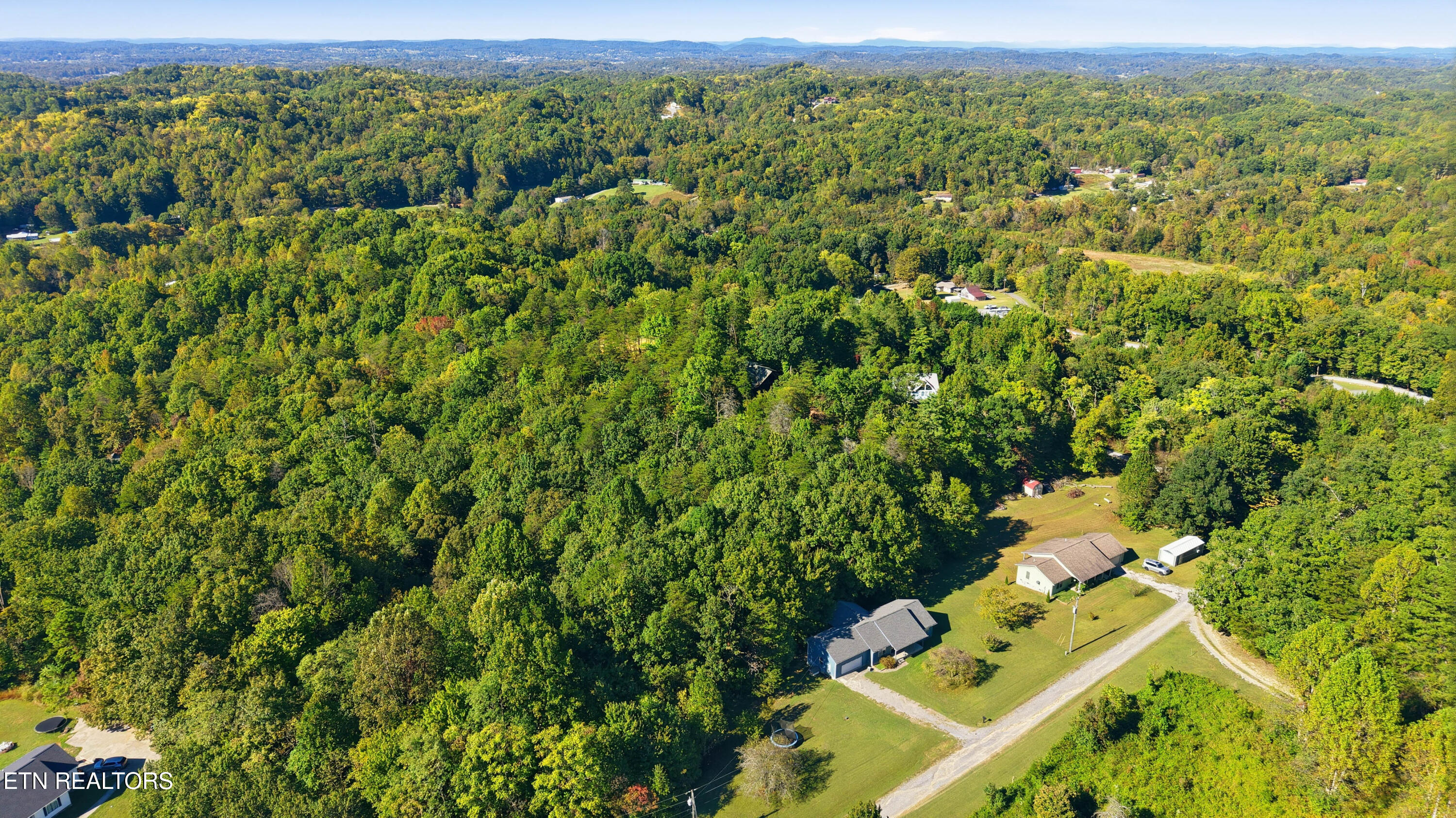Aerial view of wooded rural land with scattered homes, a gravel road, and distant ridgelines.