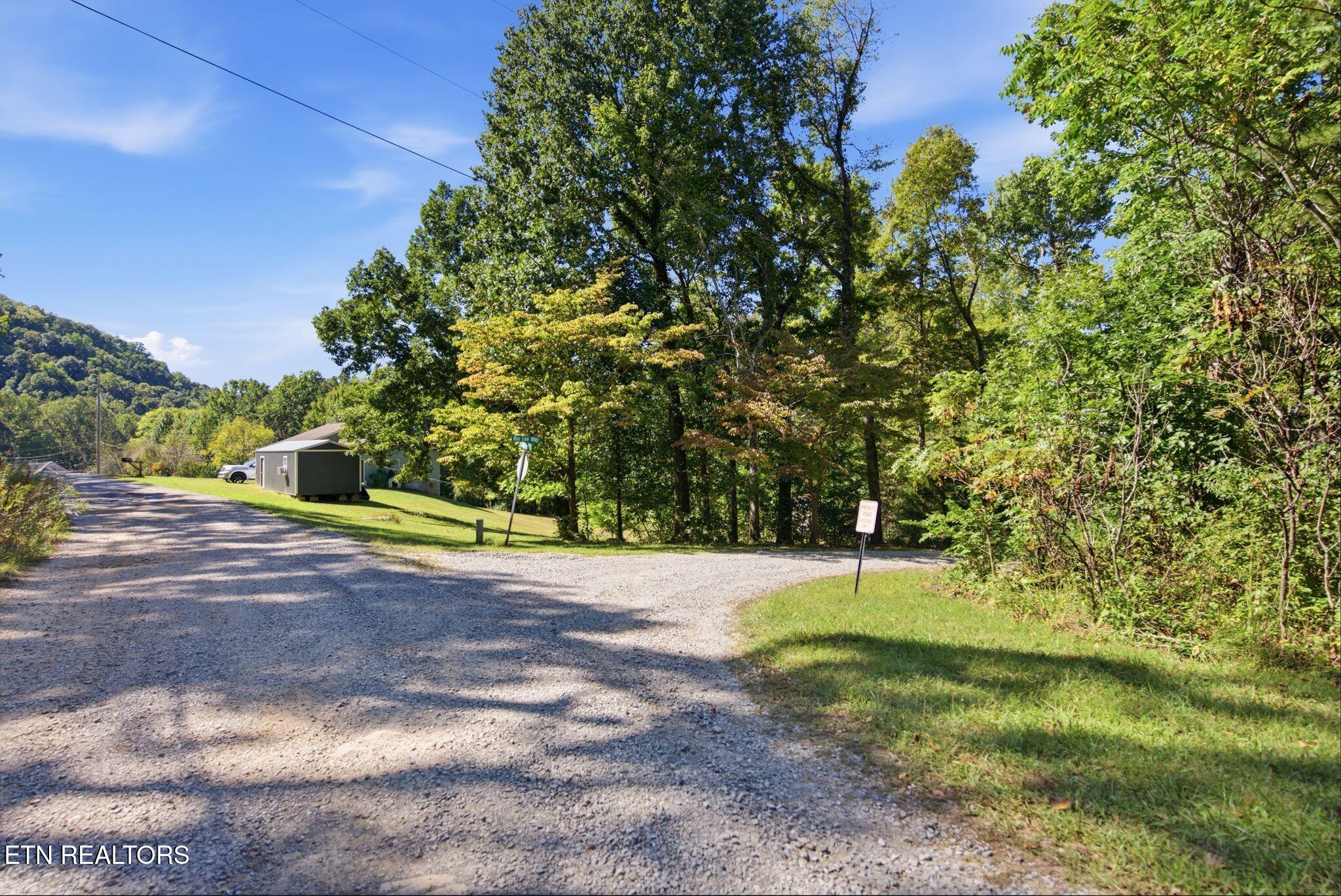 Gravel road curves beside a wooded property entrance, with a small outbuilding and wooded hills beyond.