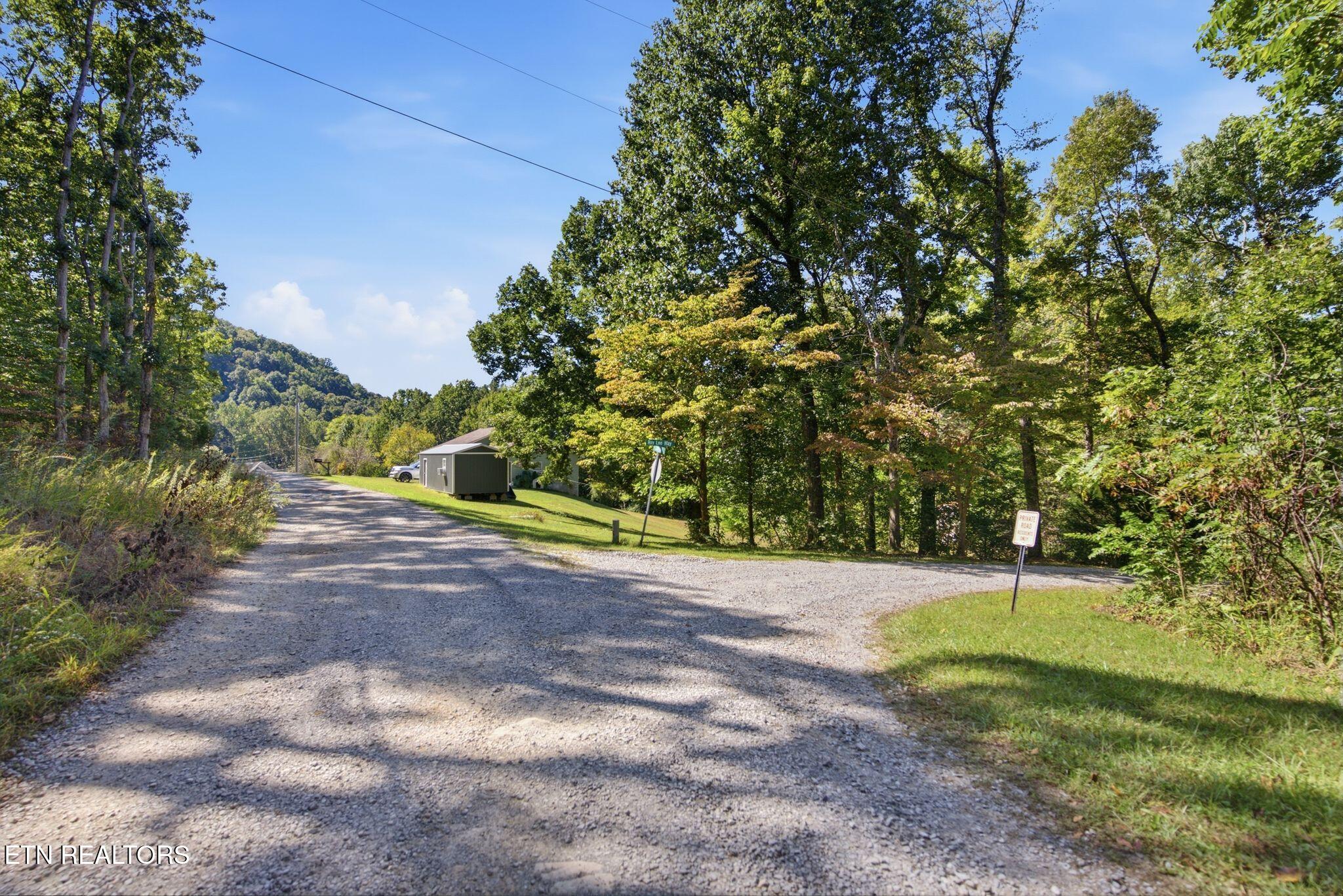 Gravel road winds past a small outbuilding through wooded land, with a forested ridge in the distance.