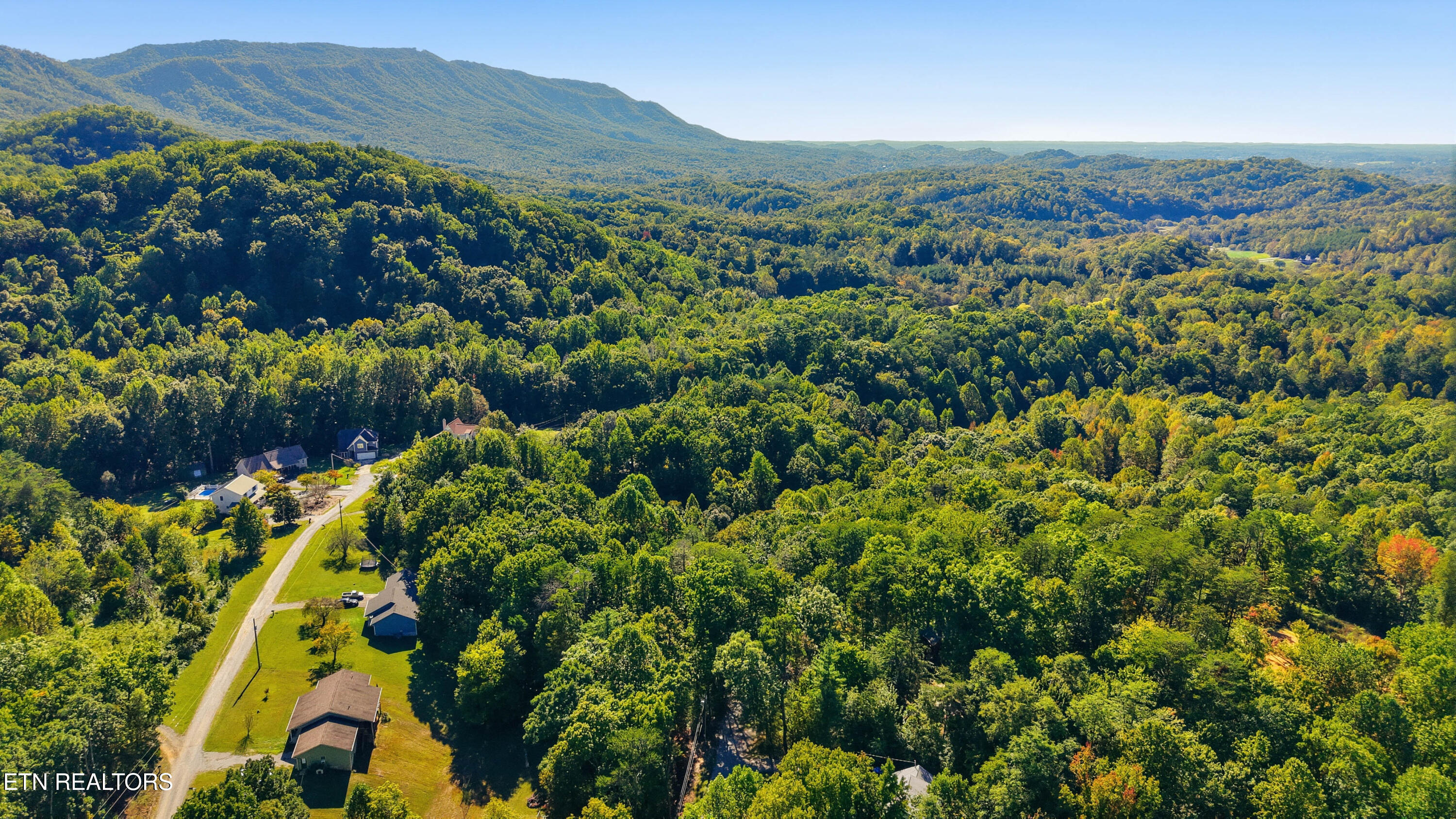 Aerial view of wooded acreage and scattered homes, with a winding road and distant mountain ridges.
