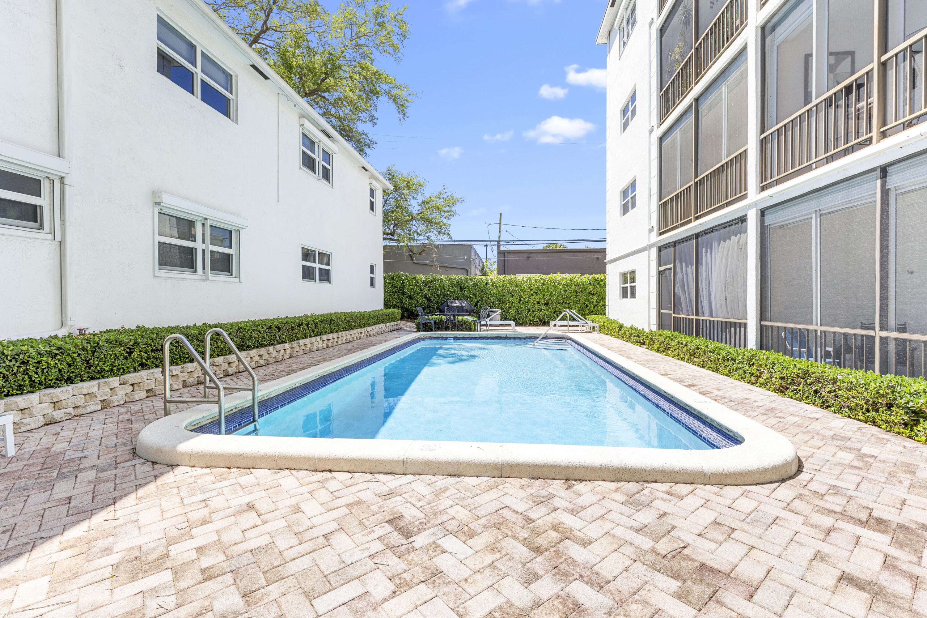 Courtyard swimming pool between white buildings, with a brick-paver deck and hedge-lined edges.