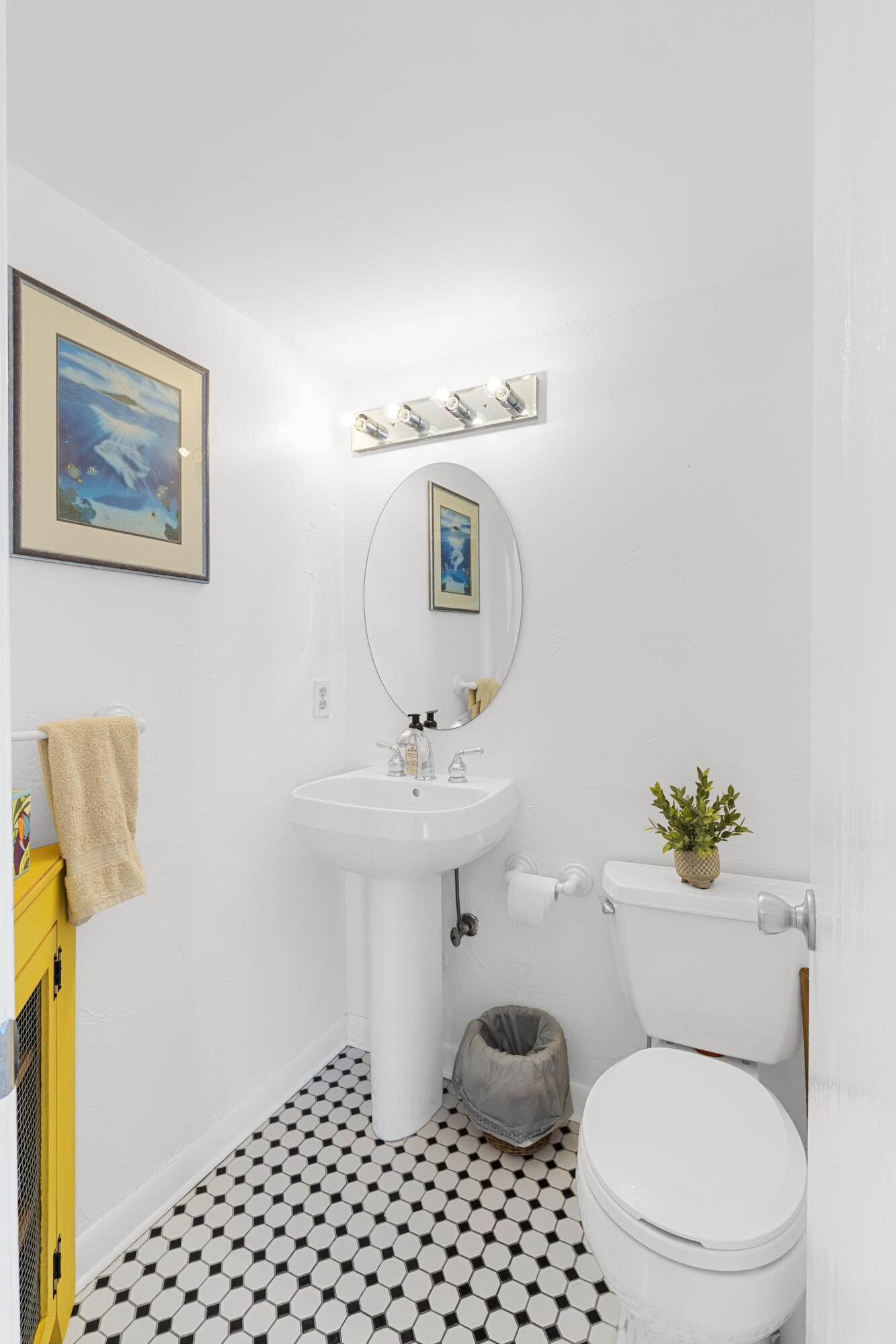 Bright bathroom with a pedestal sink, oval mirror, toilet, and black-and-white patterned floor