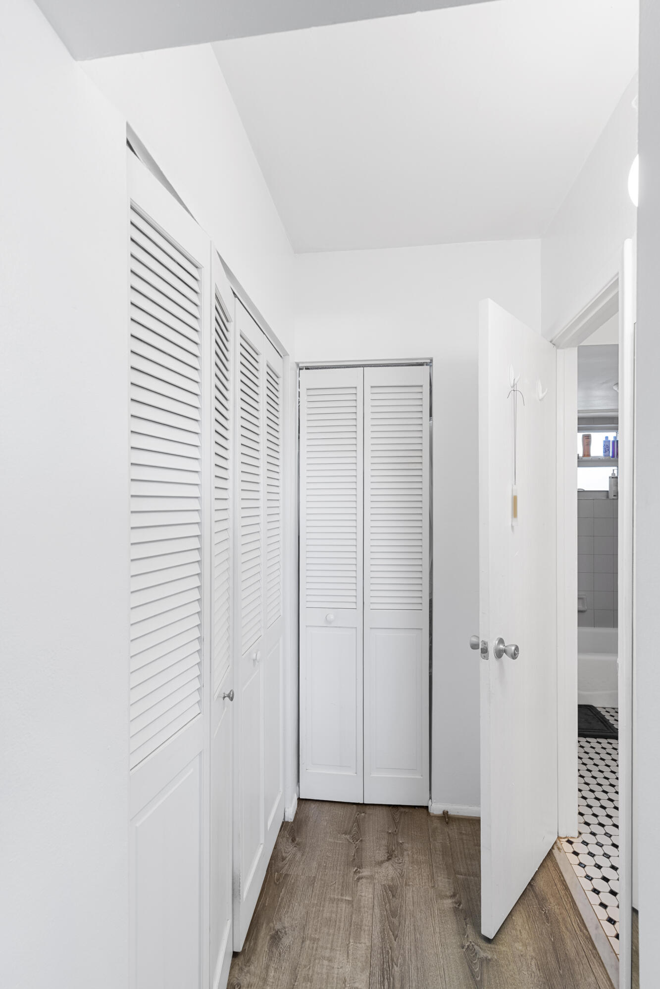 White hallway lined with louvered closet doors, opening to a bathroom with black-and-white hexagonal tile.
