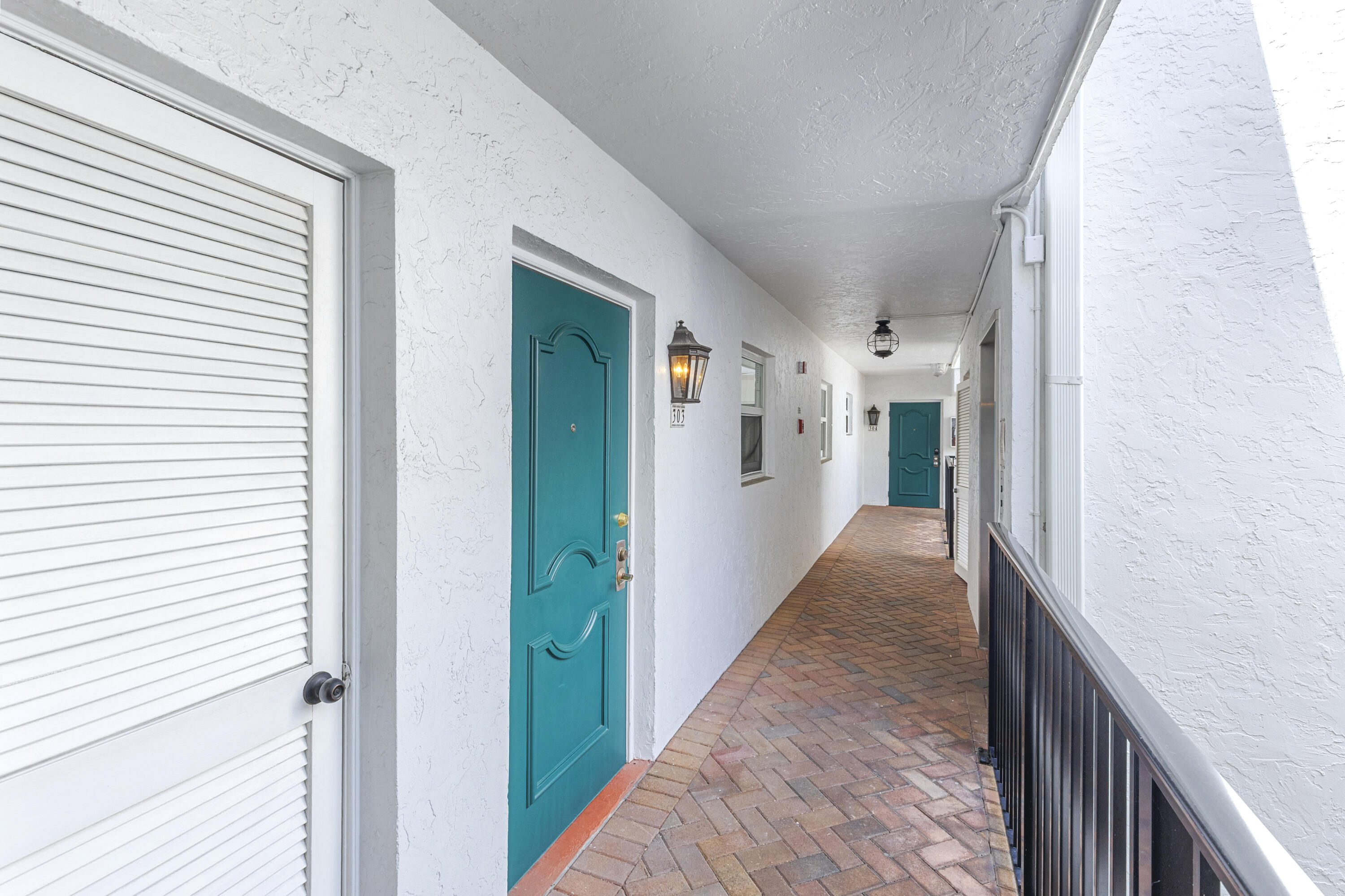 Apartment corridor with turquoise doors, a brick-paver walkway, and black railing.