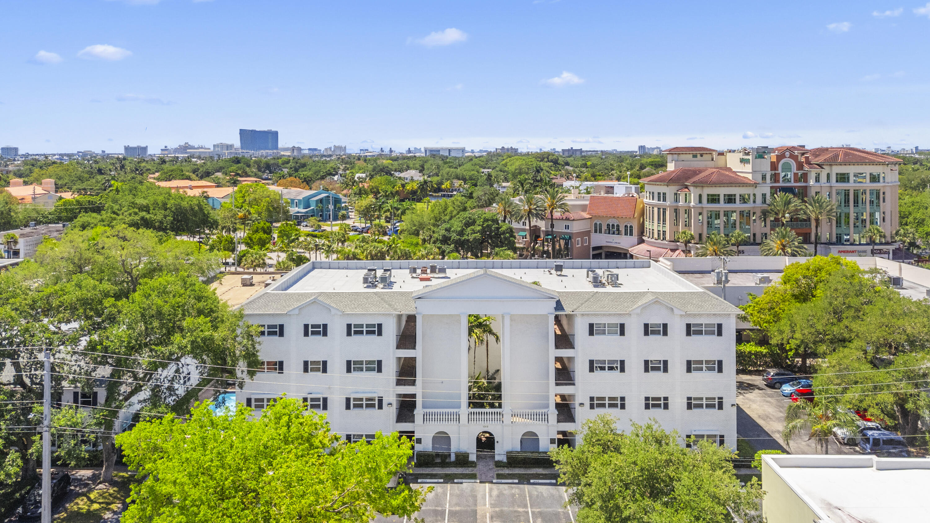 Aerial view of a white condominium building with a central entrance, trees, and a distant city skyline.