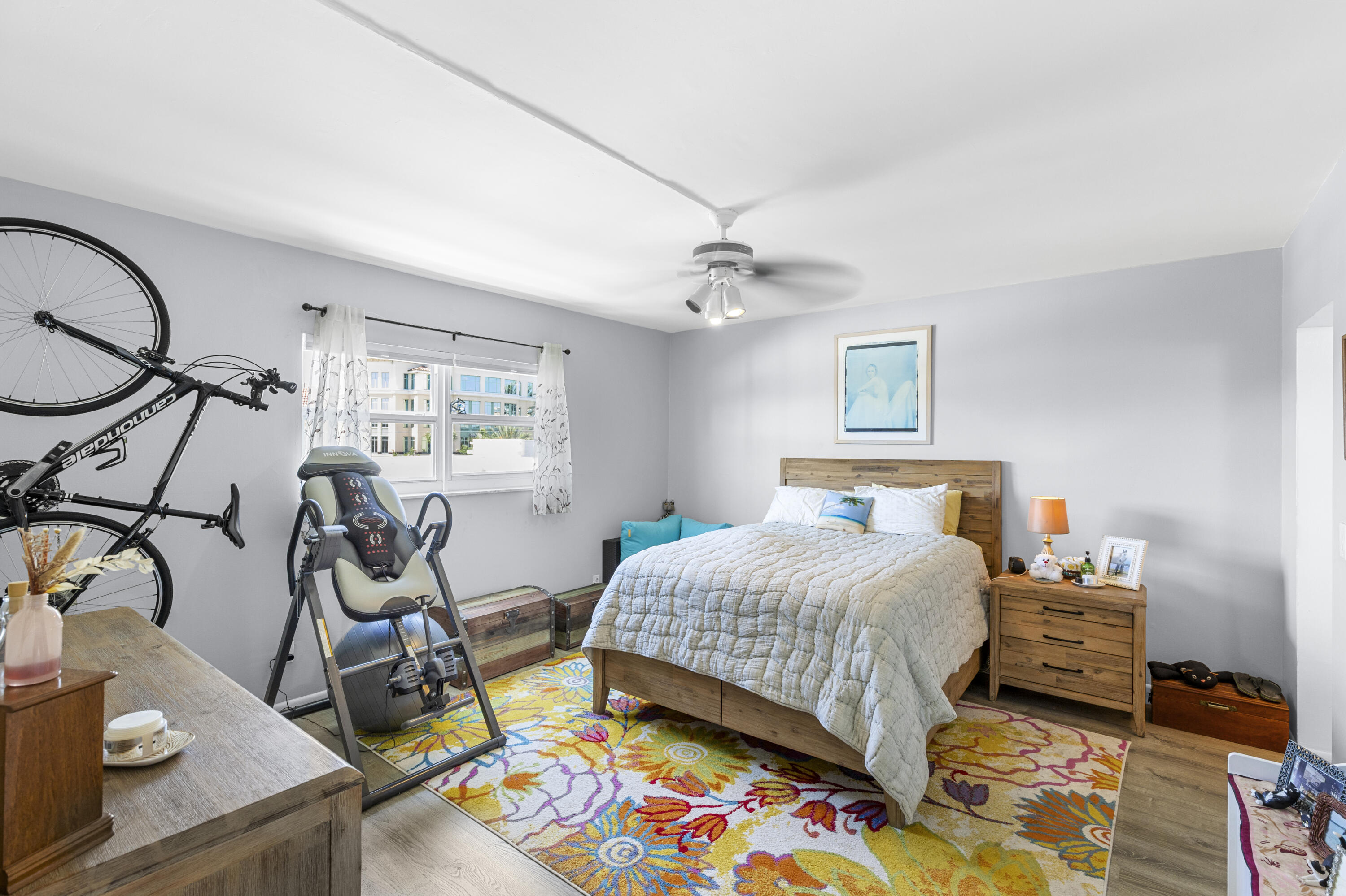 Bedroom with a wooden bed, colorful floral rug, bicycle, and inversion table beside the window.