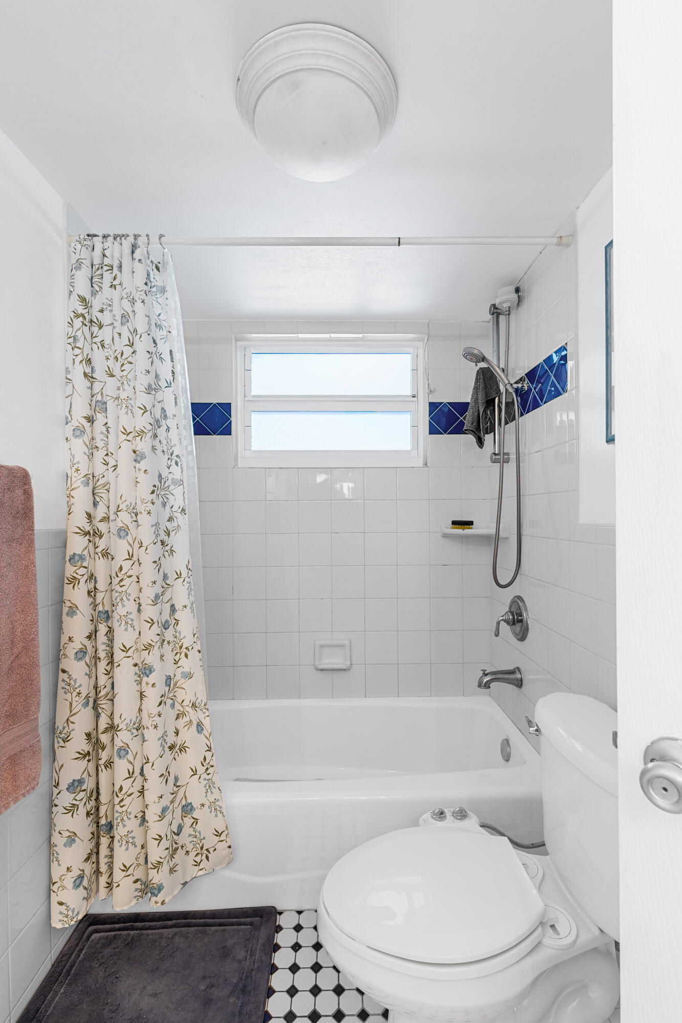 Bathroom with a floral shower curtain, blue tile accents, and a bathtub beneath a small window.