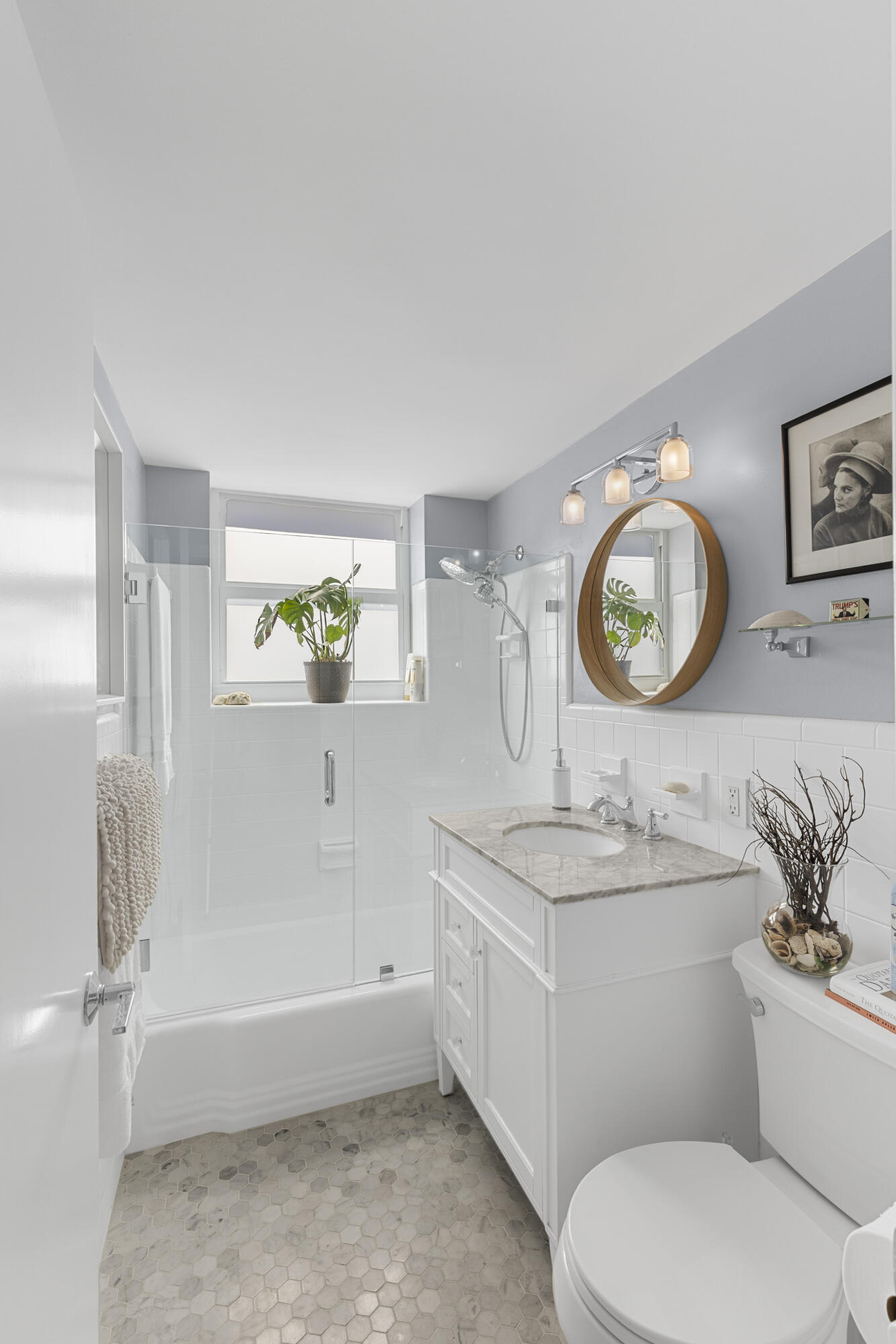 Bright bathroom with glass-enclosed tub and shower, marble vanity, and round wood-framed mirror.