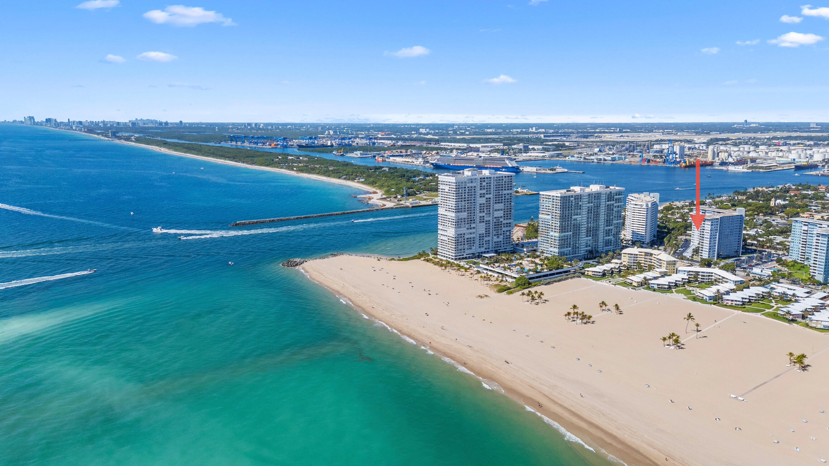 Aerial view of a beachfront condominium tower marked by a red arrow, beside turquoise water and a wide sandy beach.