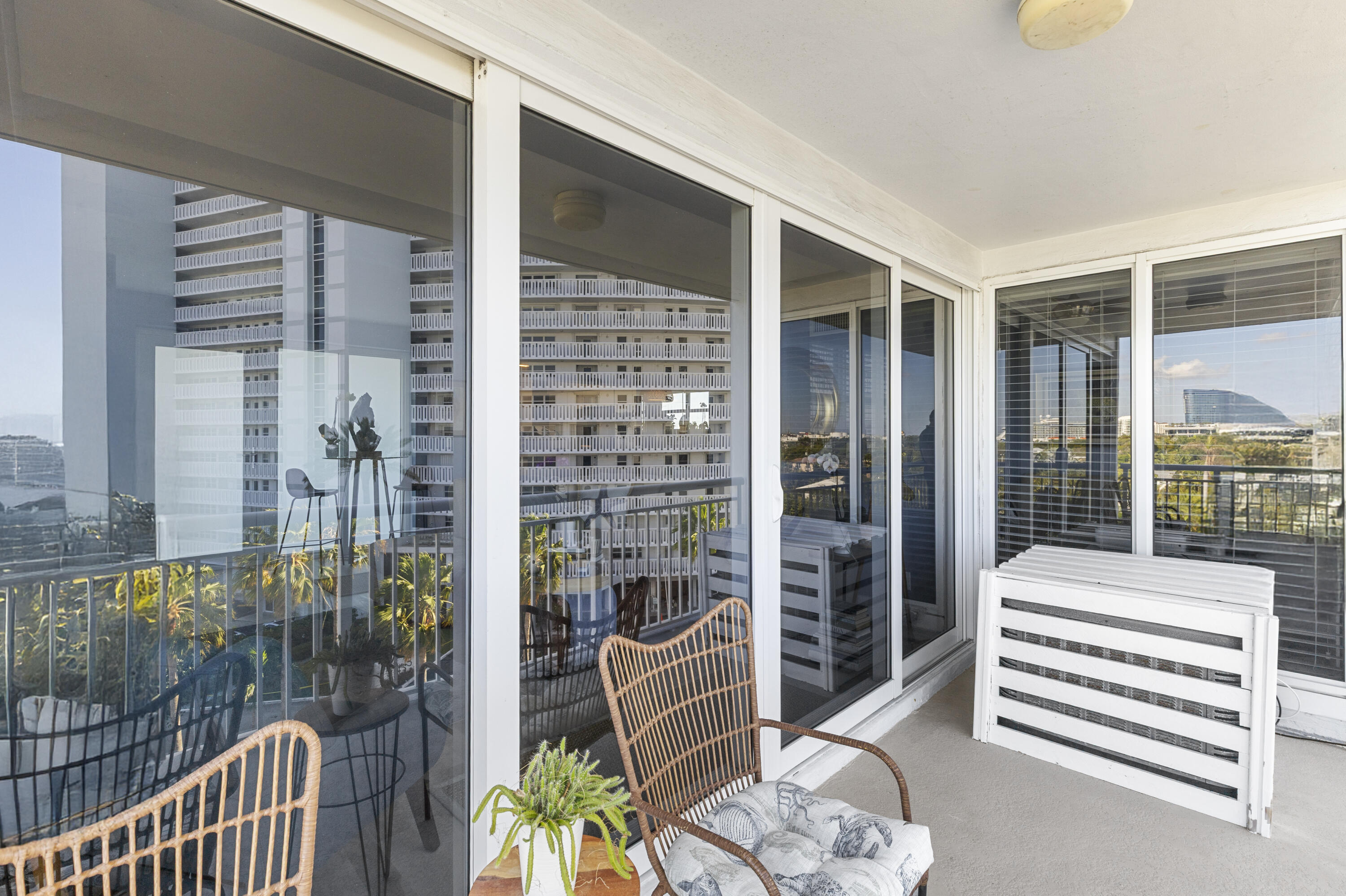 Enclosed condo balcony with wicker chairs, a potted plant, and views of nearby high-rise buildings.