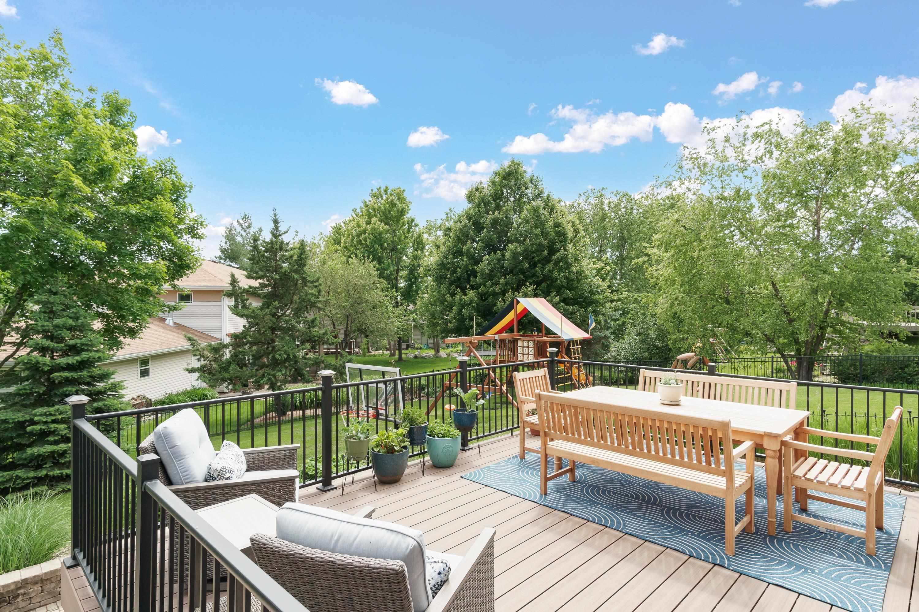 Backyard deck with a wooden dining set, lounge chairs, and a colorful children’s playset beyond the railing.