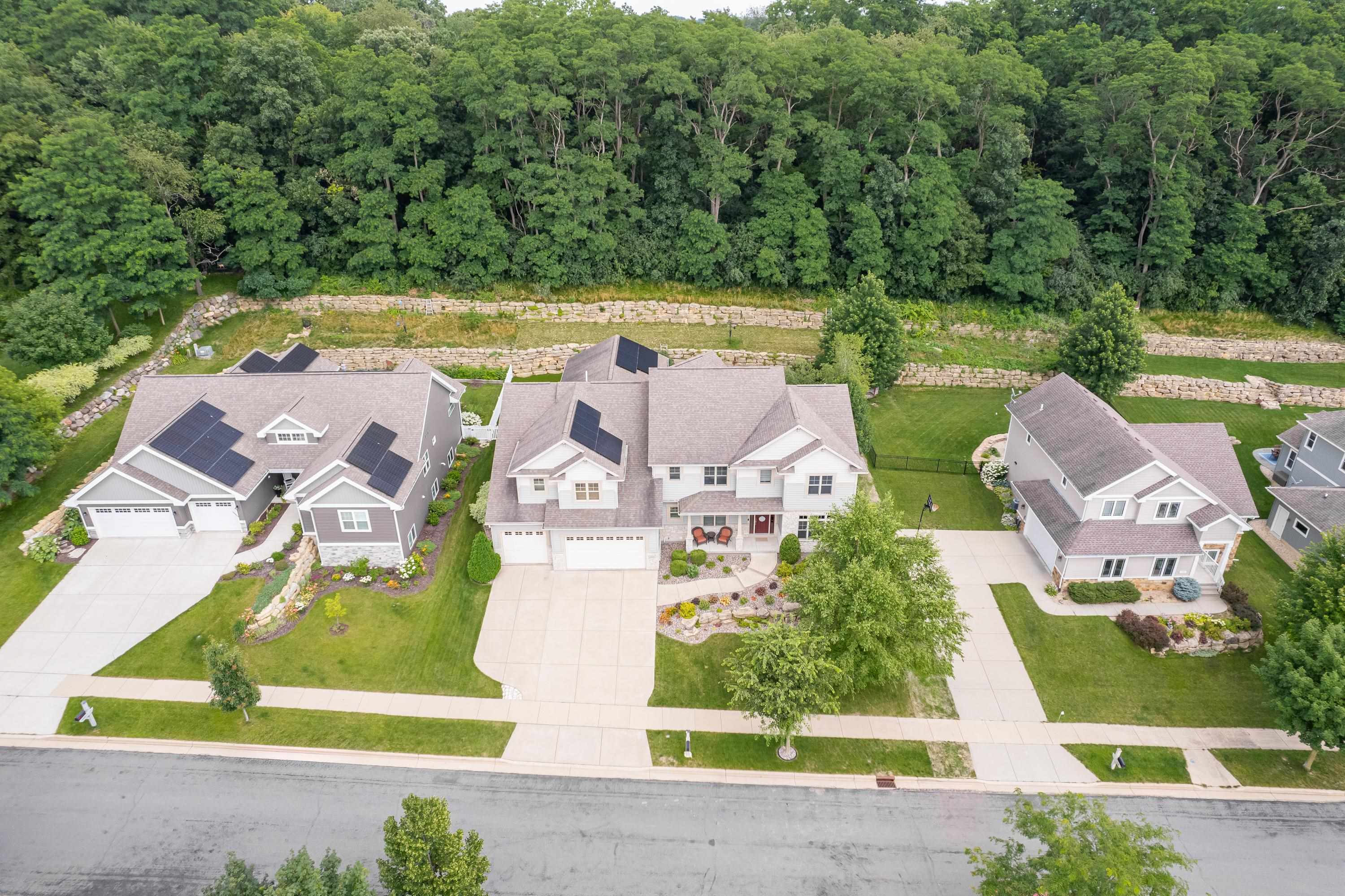 Aerial view of suburban homes beside dense woods, with solar panels and stone retaining walls.