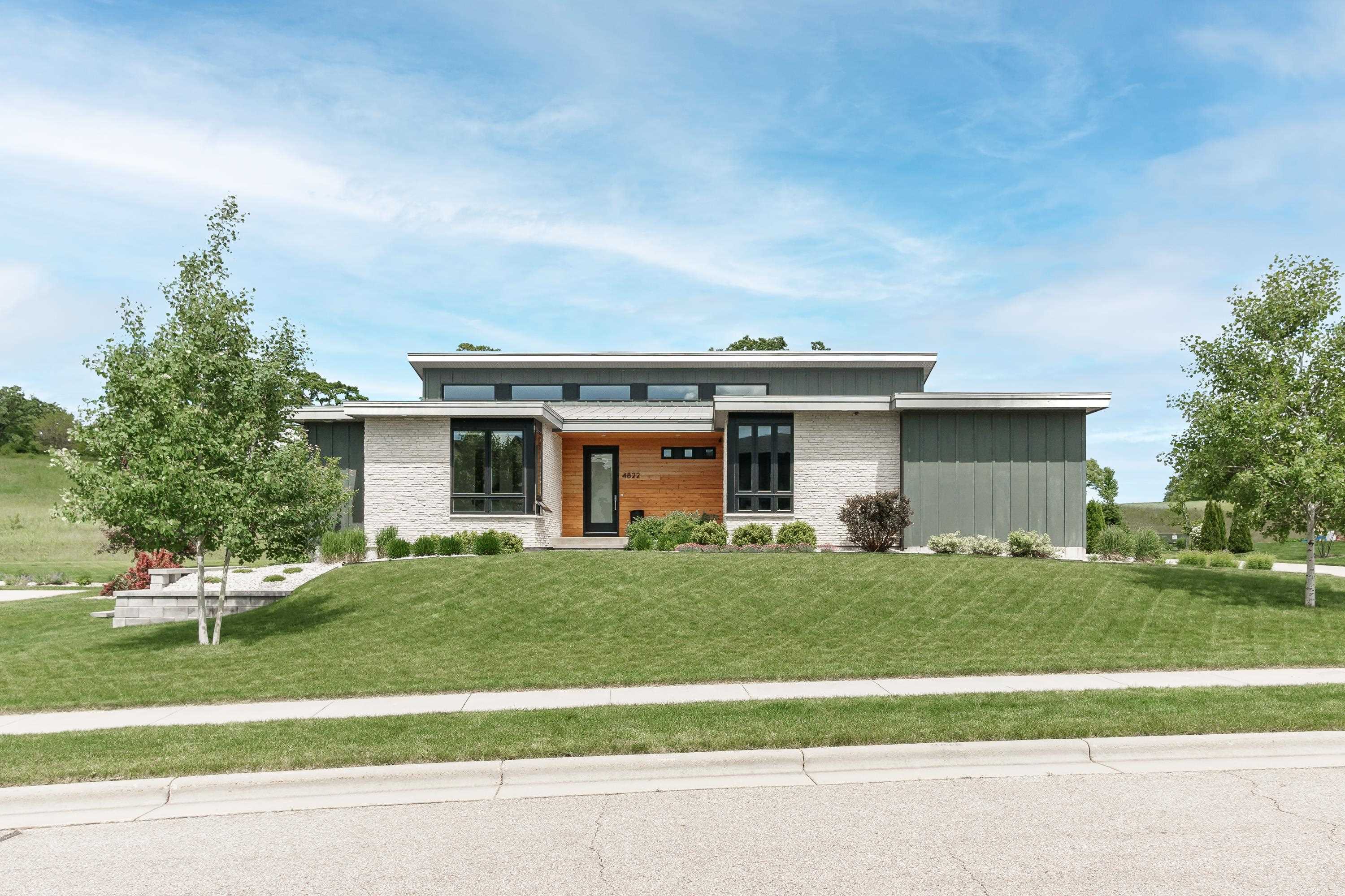 Modern home with white brick, dark siding, and a wood-framed entrance, set behind a broad green lawn.
