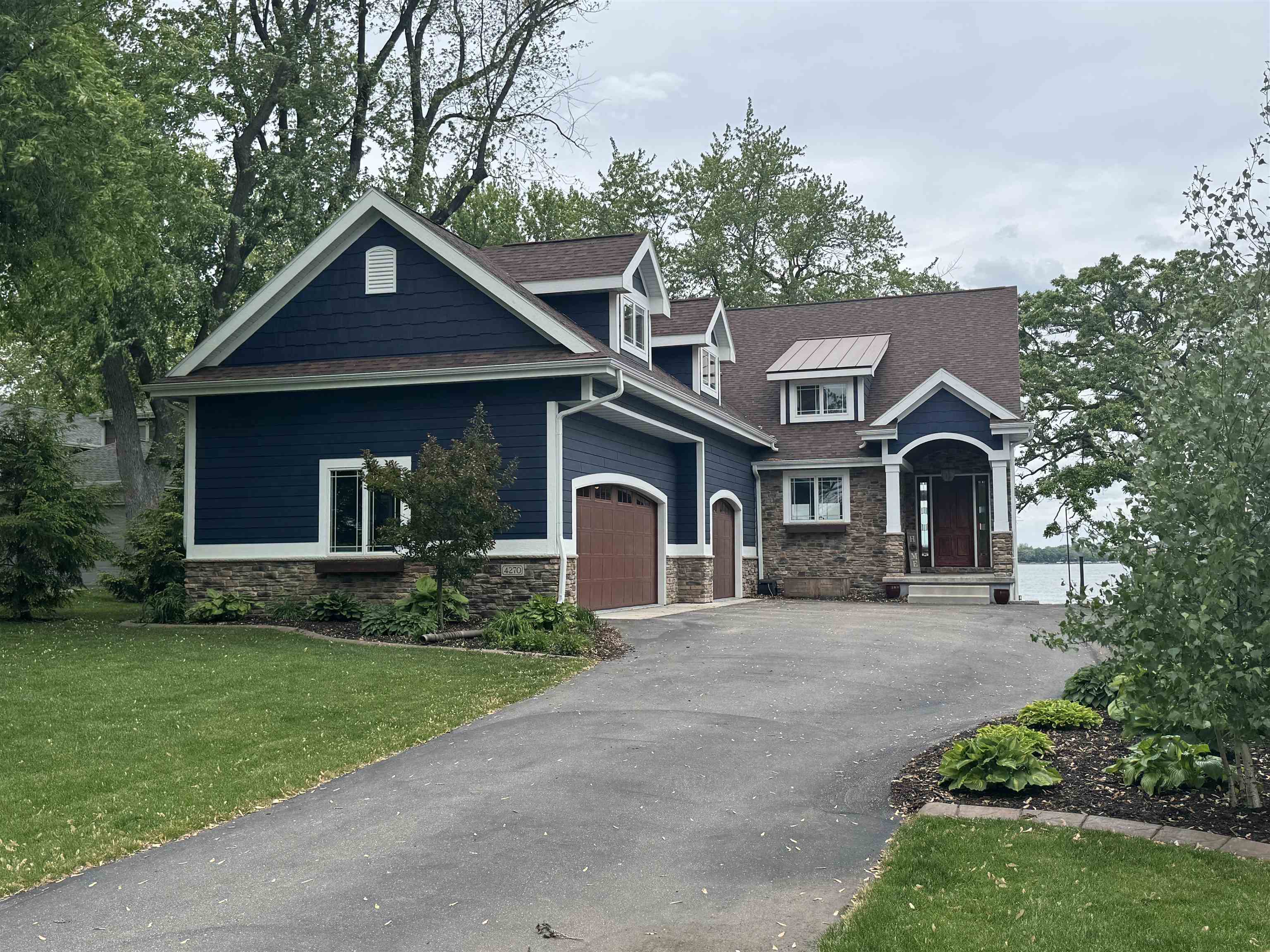 Two-story navy-blue and stone home with a broad driveway, mature trees, and water beside it.
