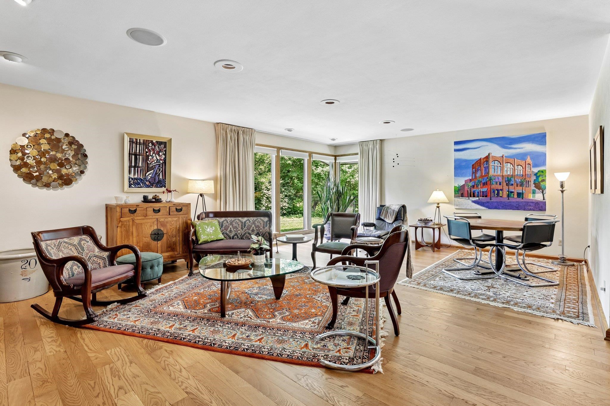 Open-plan living and dining room with hardwood floors, patterned rugs, and large windows overlooking greenery.