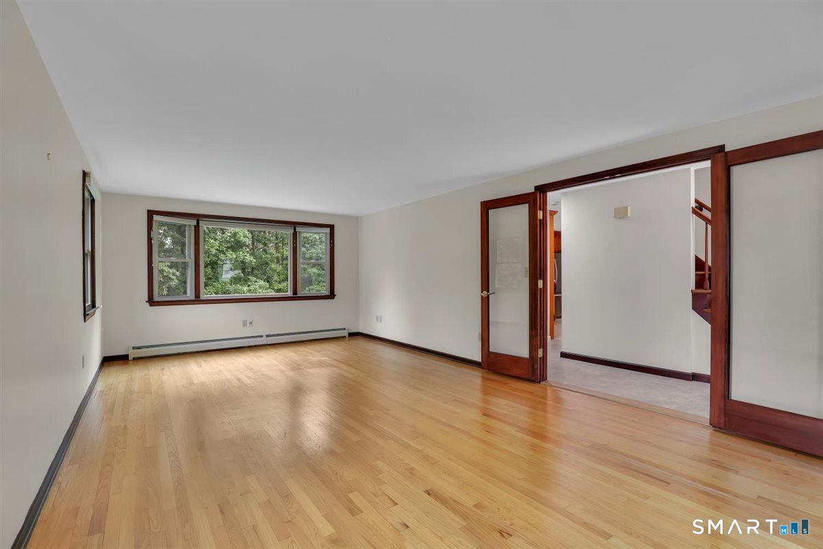 Empty living room with hardwood floors, a wide window overlooking trees, and dark wood trim.