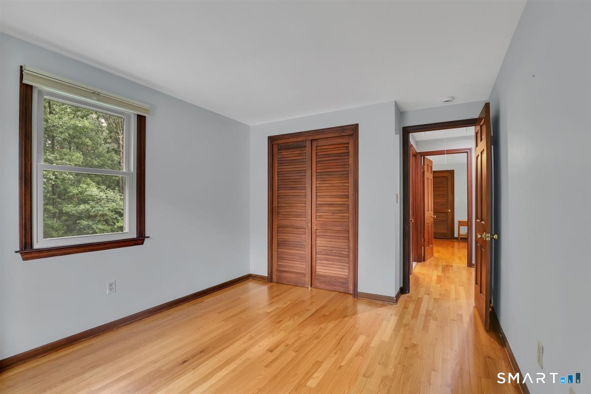 Unfurnished bedroom with hardwood floors, louvered closet doors, and a window overlooking trees.