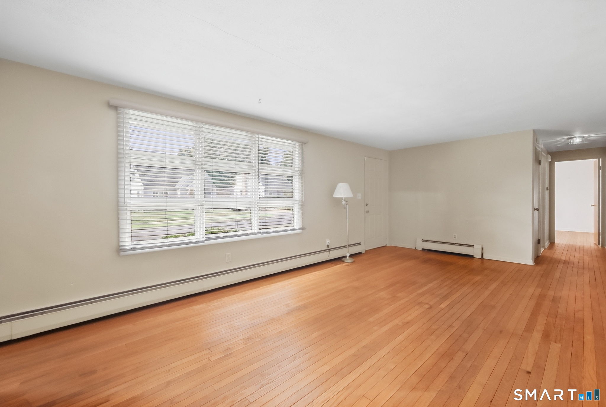 Living room with hardwood floors, wide window with white blinds, and baseboard heating.