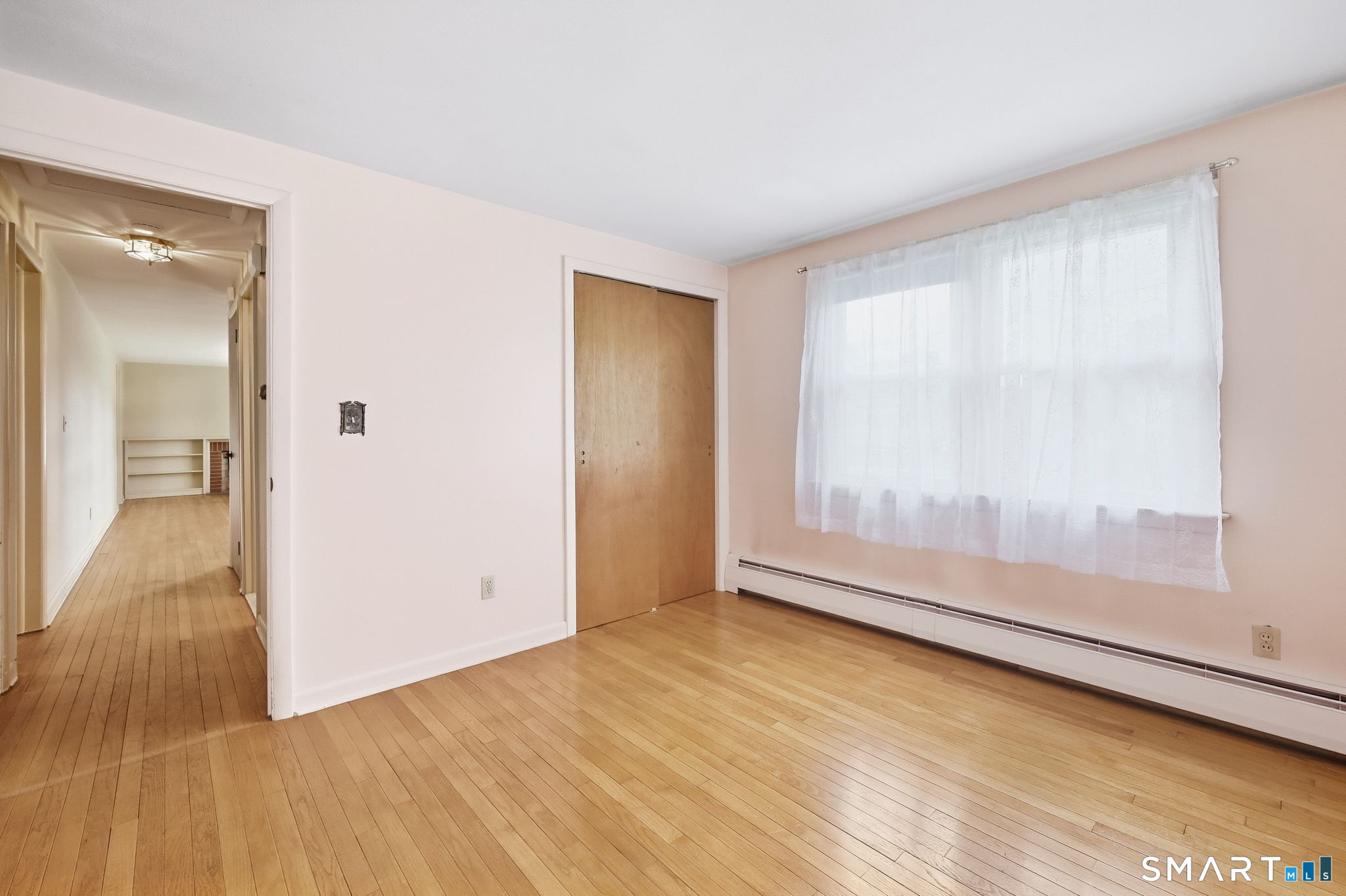 Empty room with hardwood floors, a large curtained window, and a sliding wooden closet door.