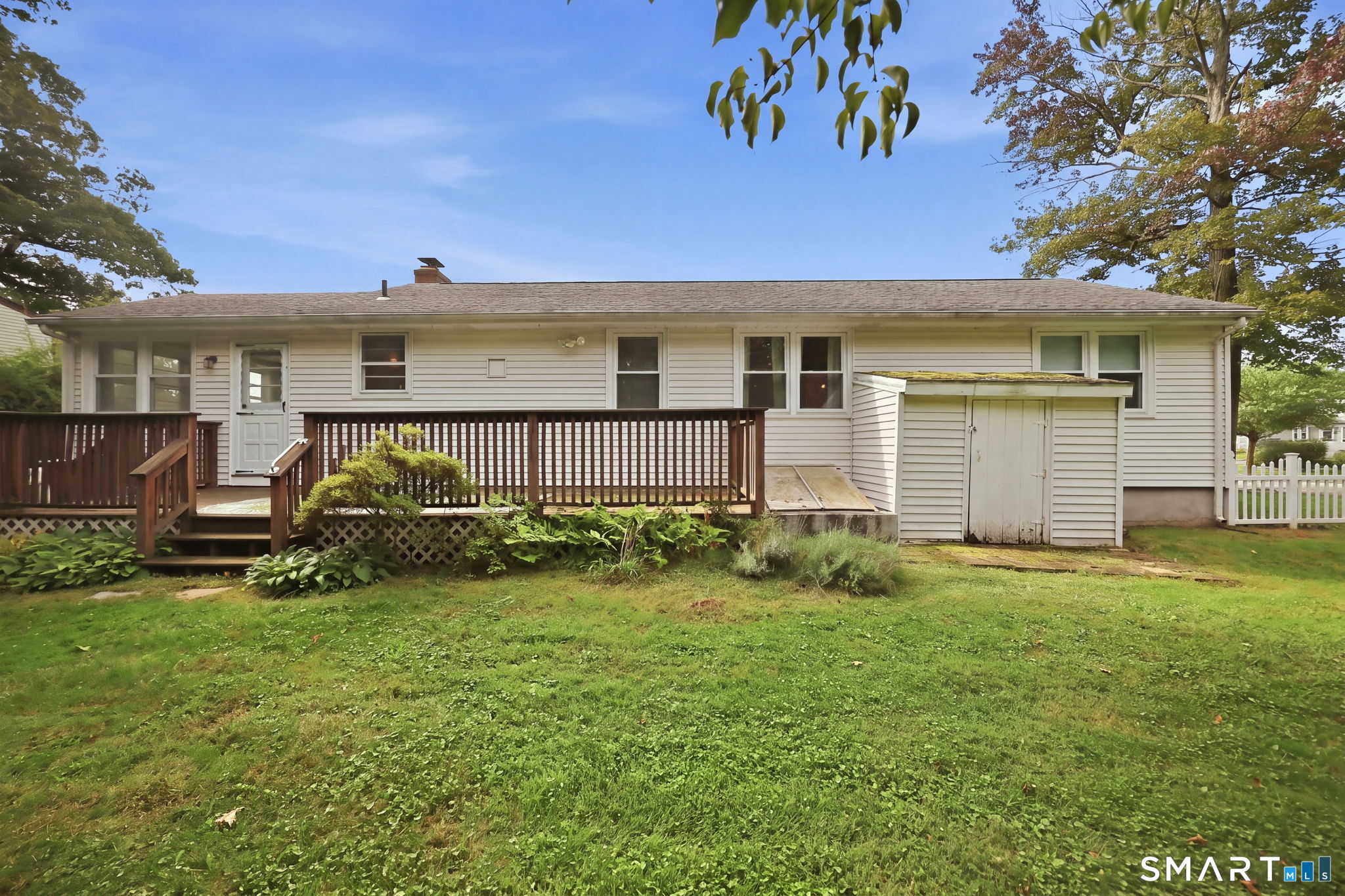 Rear of a single-story house with a long wooden deck, attached shed, and broad lawn.