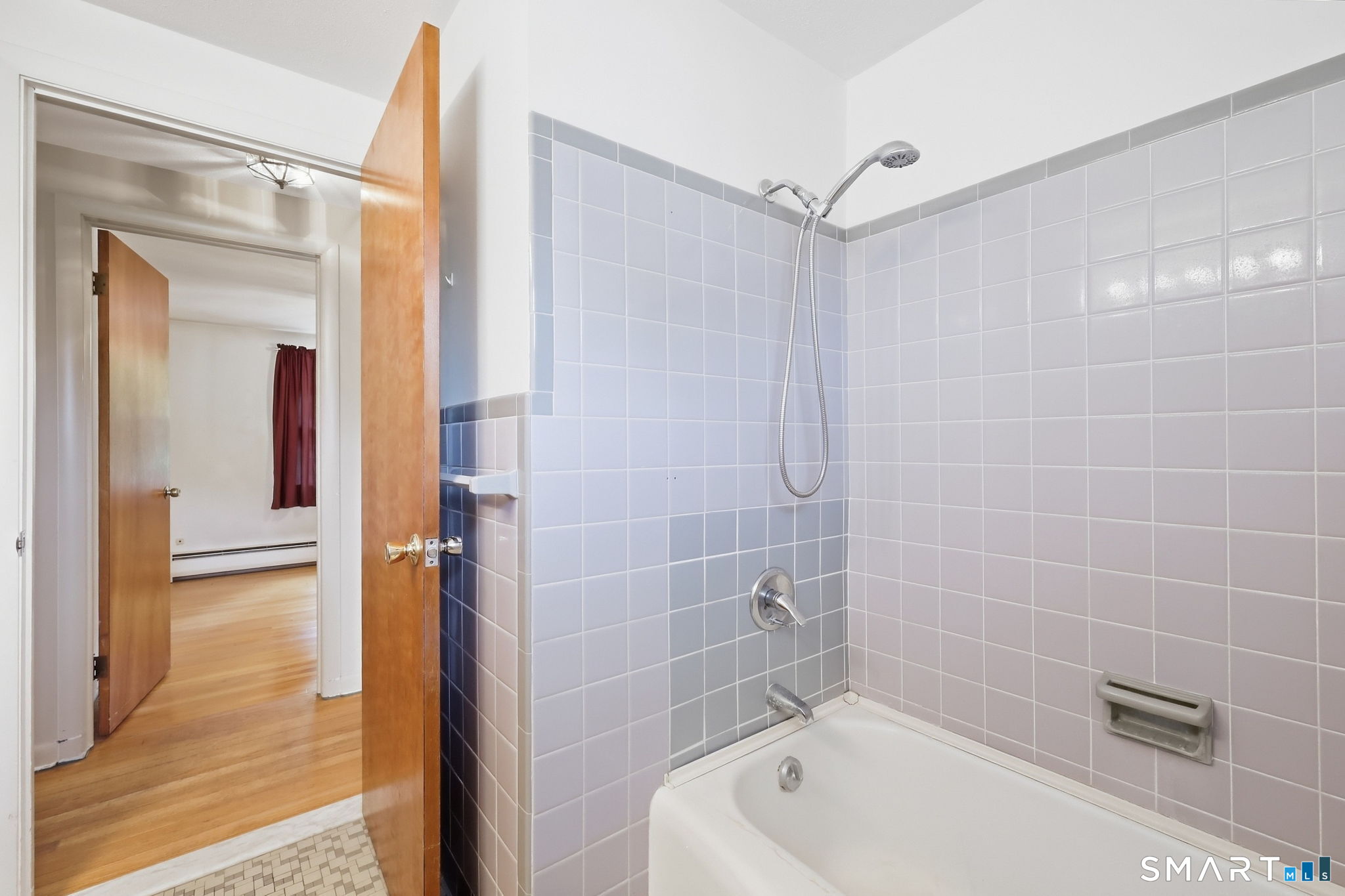 Bathroom with a white tub, pale tiled shower walls, and an open wooden door to a hardwood-floored room.