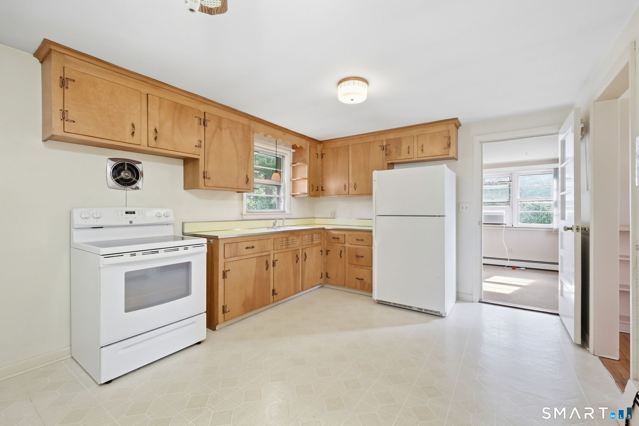 Kitchen with honey-colored wood cabinets, white appliances, and a doorway to a sunlit room.